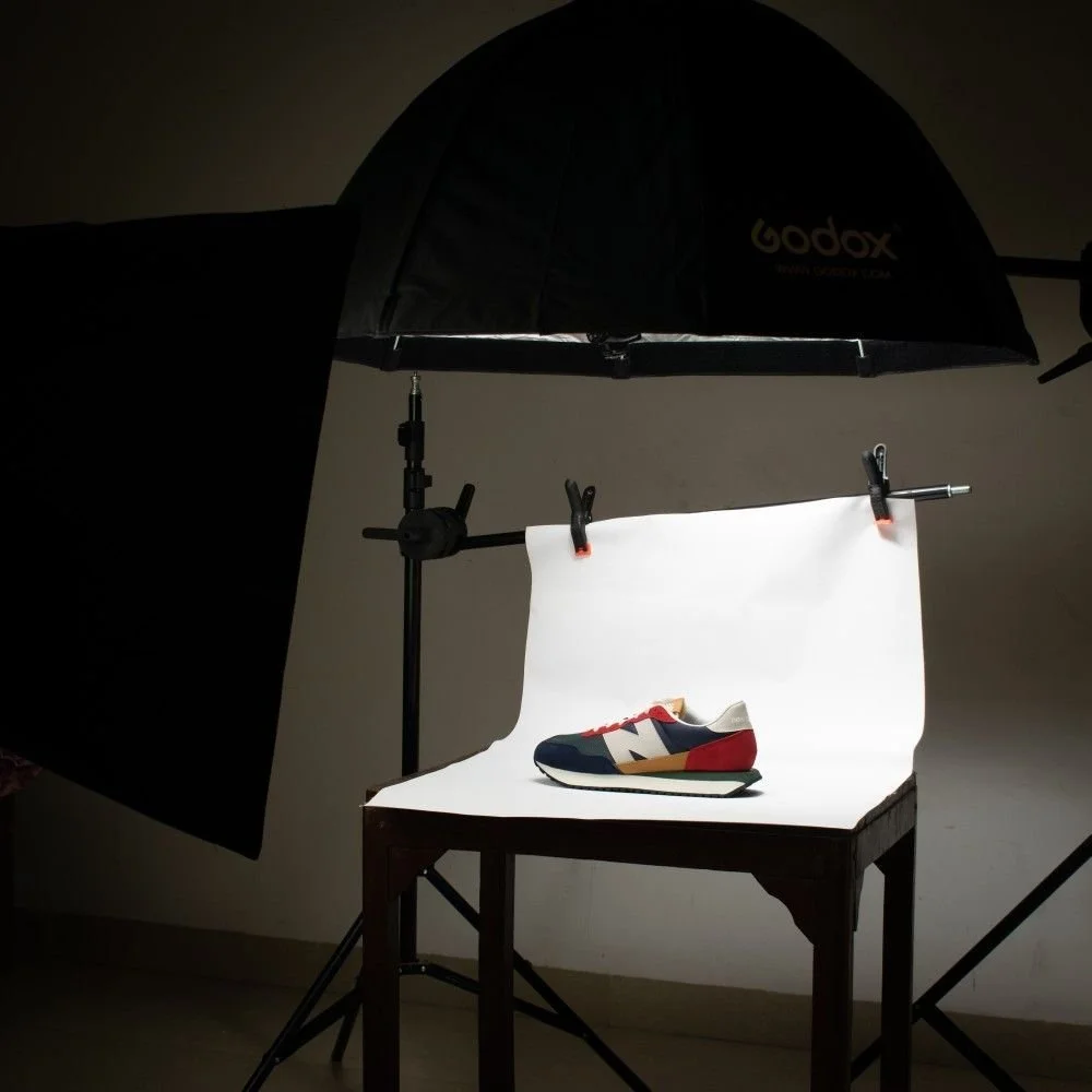 Material de fotografía con una especie de estudio, que incluye un paraguas difusor, luces, una mesa y ropa, con un zapato de diseñador colorido en un fondo blanco.