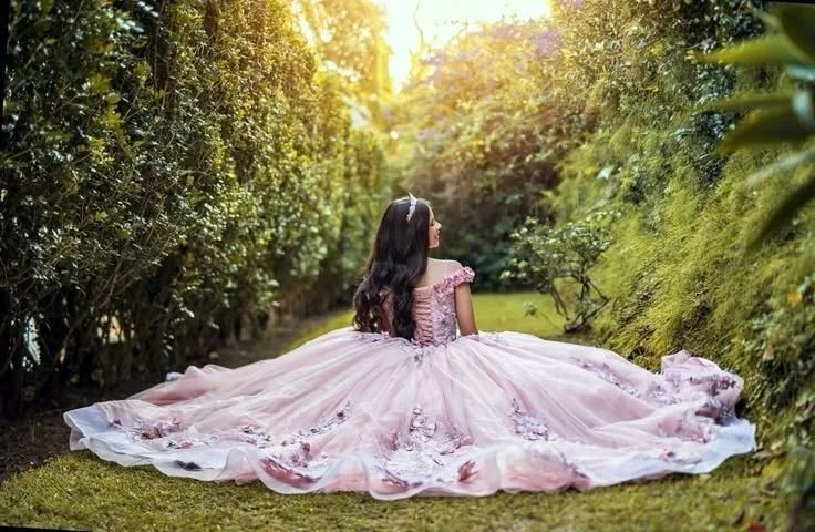 Una joven con vestido rosa largo y en un entorno natural, sentada sobre la hierba en un camino entre arbustos verdes, con el sol brillando detrás.