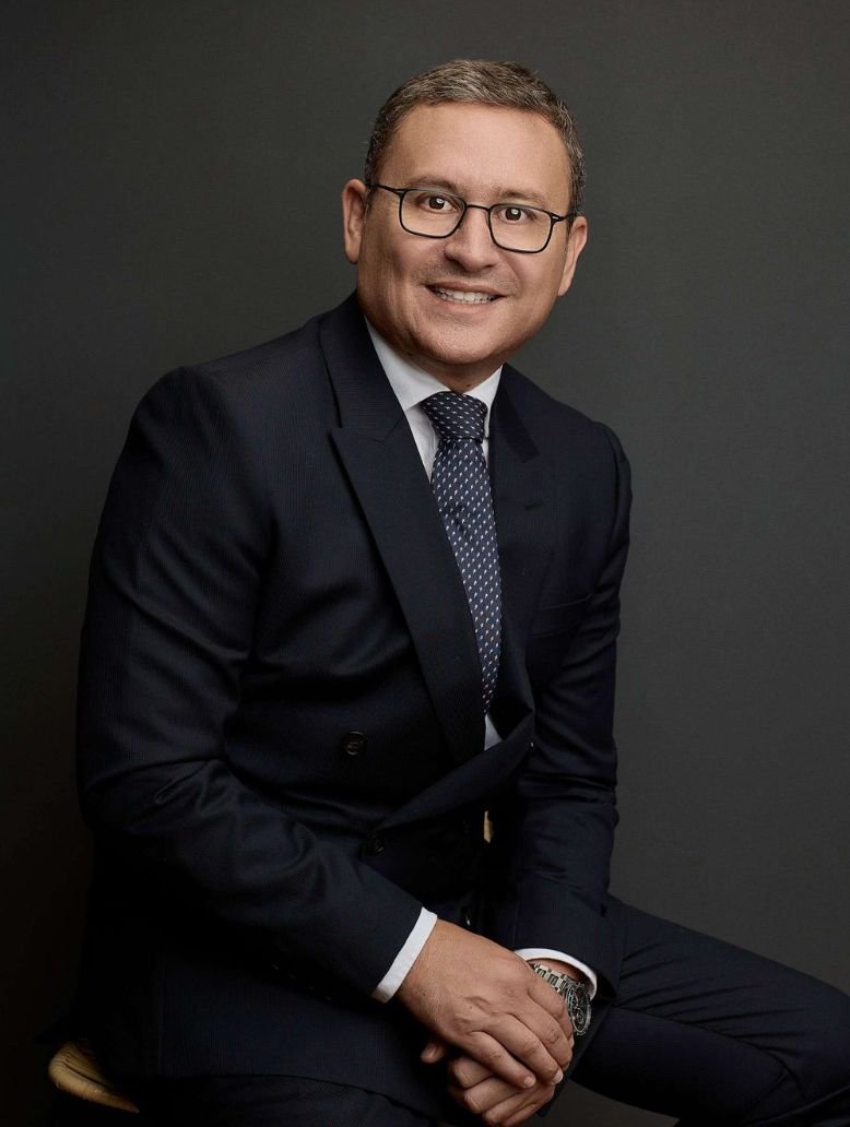Hombre con traje oscuro, camiseta blanca, y gafas, sentado frente a un fondo gris oscuro, sonriendo.