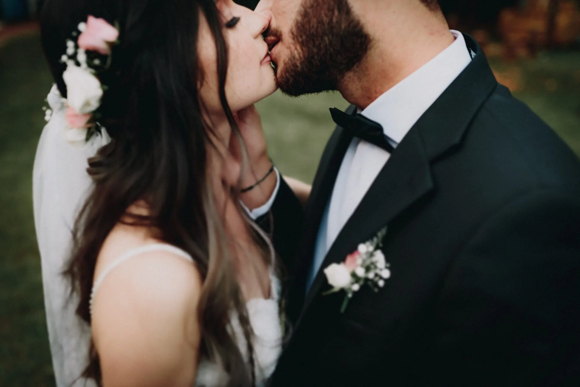 Pareja casada besándose con vestido de novia y traje de esmoquin en un ambiente exterior.