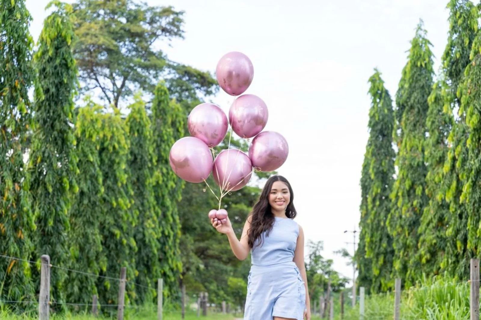 Una joven sonriente sosteniendo globos rosados en un campo rodeado de árboles verdes.