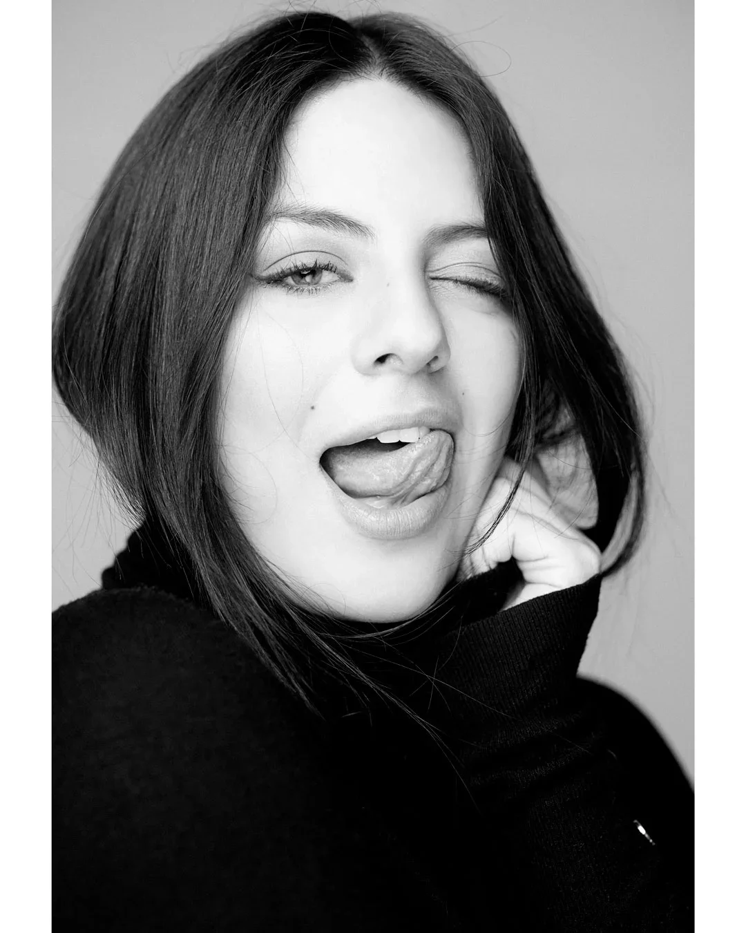 Retrato en blanco y negro de una mujer con cabello oscuro, guiñando un ojo, levantando la lengua y sonriendo parcialmente mientras se toca el cuello.