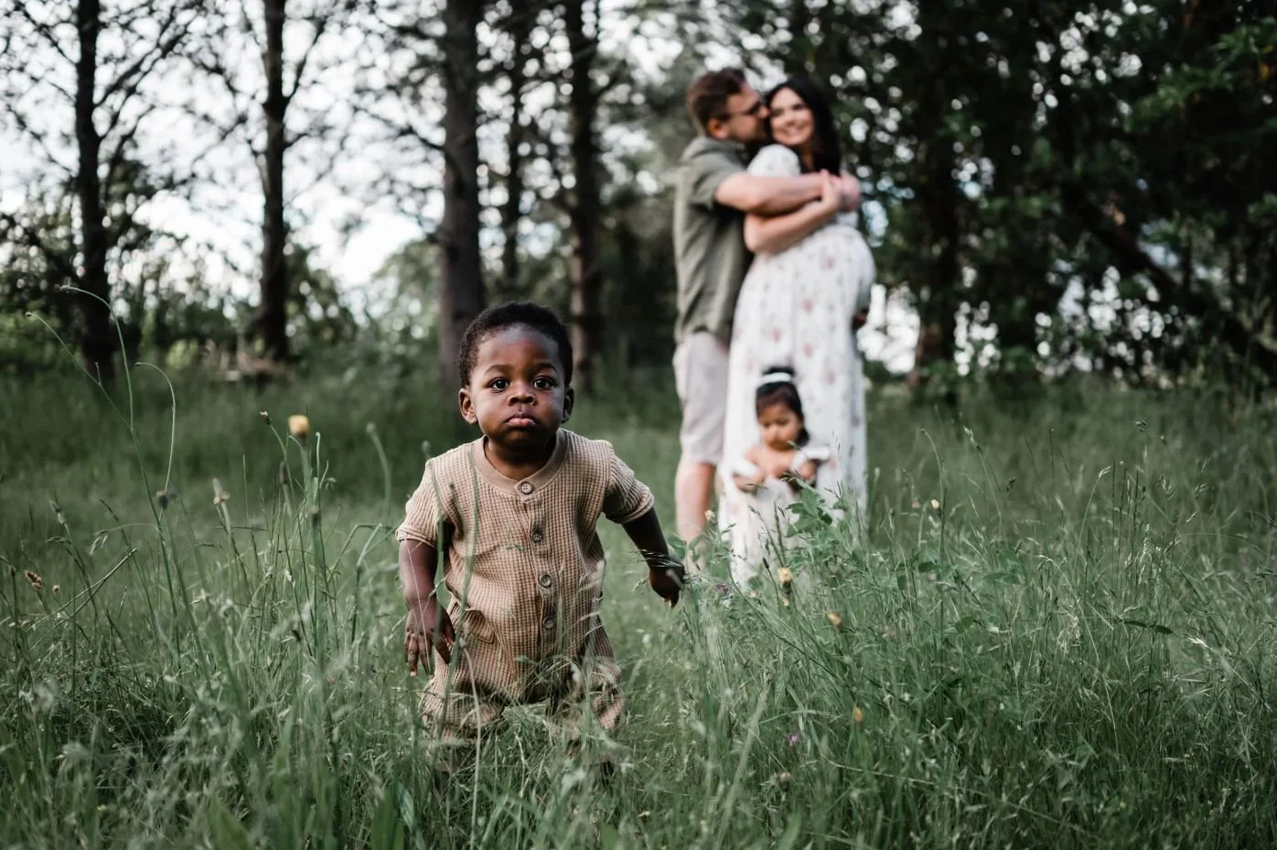 Una familia en un campo de hierba con árboles y un cielo despejado. Un niño pequeño en primer plano, con ropa beige y expresión curiosa. En el fondo, un hombre y una mujer abrazados, y una niña menor entre ellos, todos sonriendo en la naturaleza.