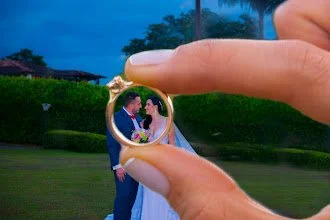 Una pareja de novios de pie en un parque, siendo vista a través de un anillo de oro que alguien sostiene con los dedos estirados, durante el atardecer.