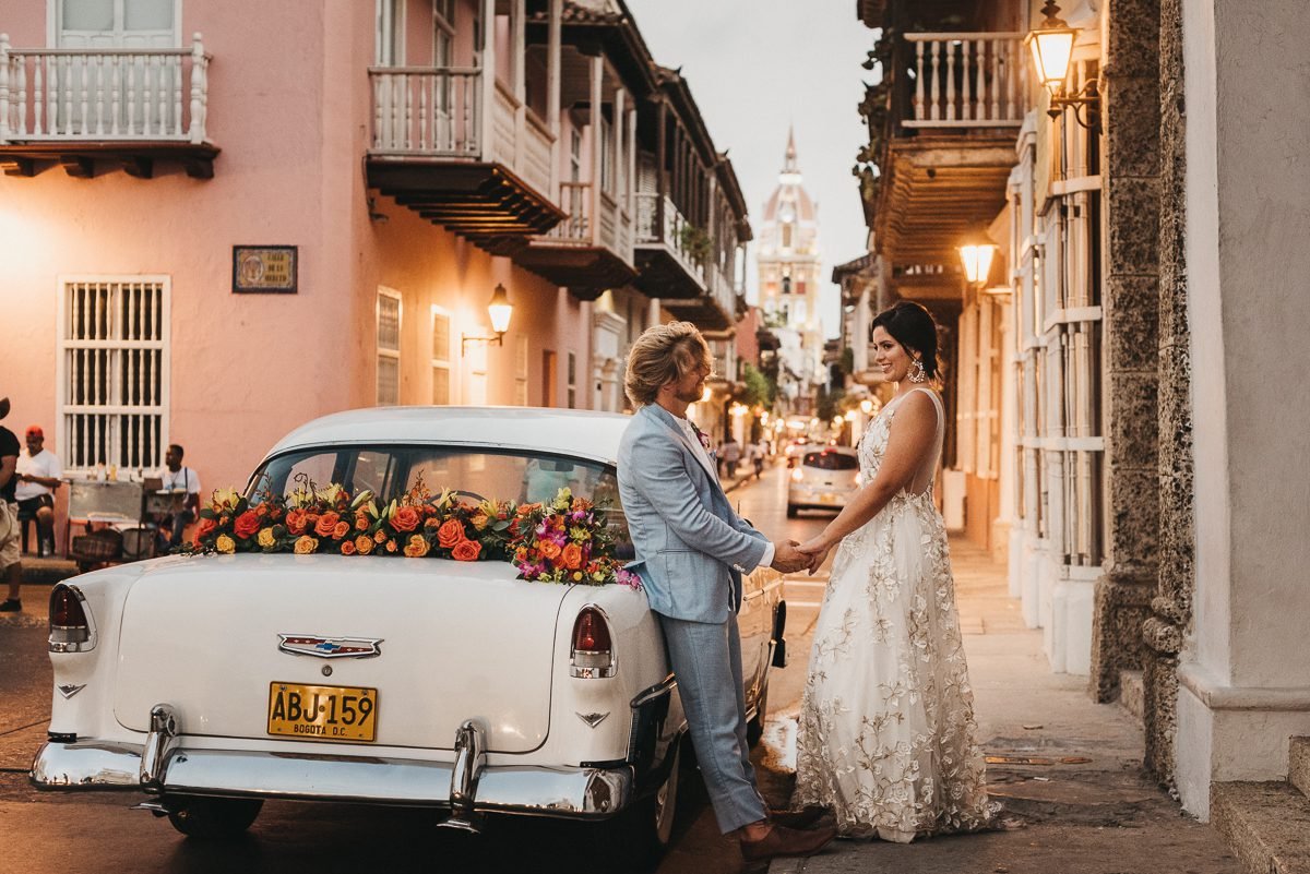 Pareja de novios sosteniéndose de las manos frente a un coche antiguo decorado con flores durante una boda en la calle en la tarde