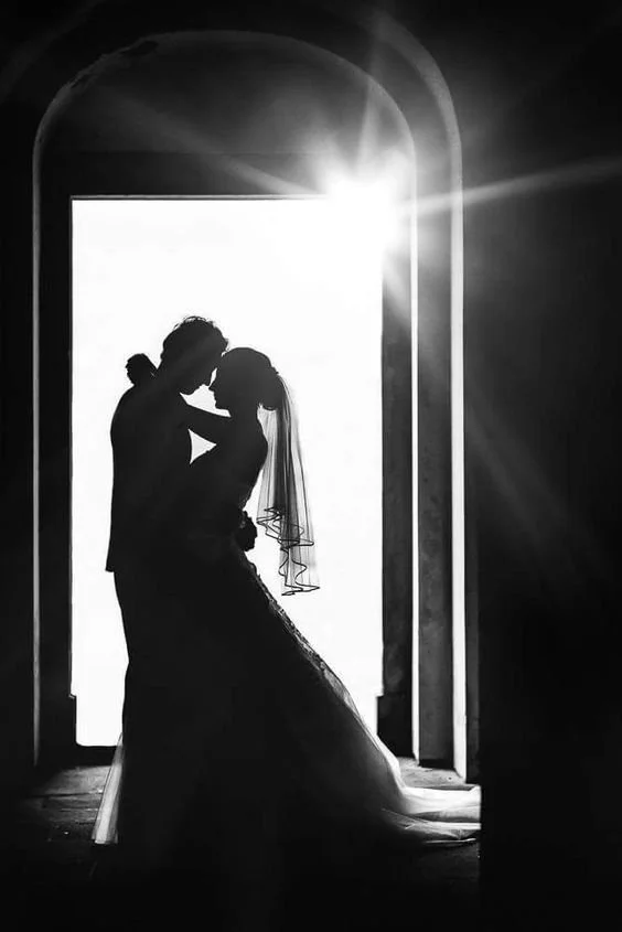 Silhueta de una pareja de novios en un abrazo romántico, con la luz del sol brillando a través de una ventana arqueada, en un entorno de interior