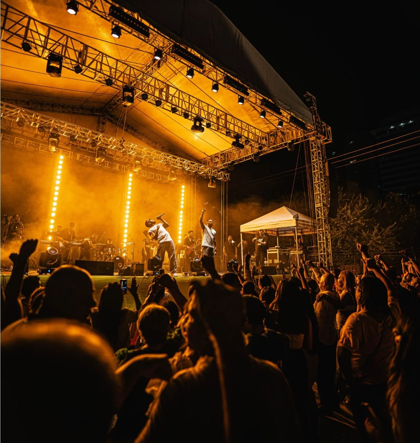 Concierto en vivo con músicos en el escenario bajo luces naranjas, público disfrutando y bailando, noche y tejado del escenario visible.