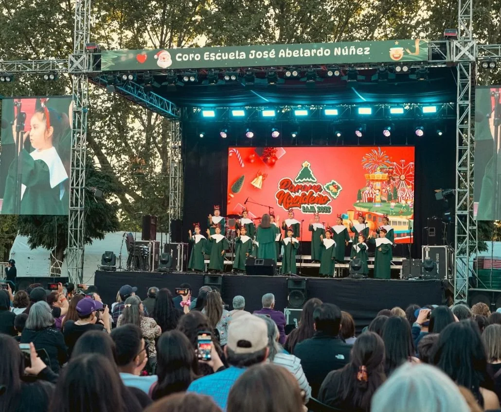 Coro escolar durante una presentación navideña en un escenario al aire libre con audiencia de adultos y niños.