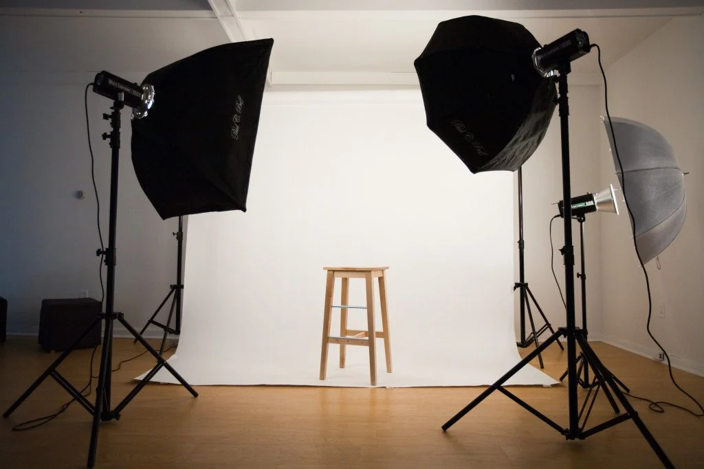 Estudio fotográfico con fondo blanco y taburete de madera en el centro, con ocho luces de estudio y difusores alrededor.