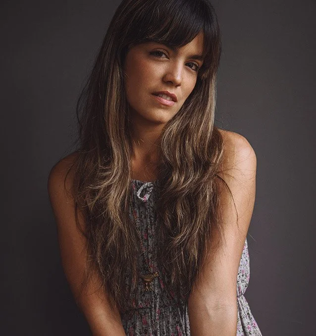 Retrato de una mujer con cabello largo y rizado, posando contra un fondo gris oscuro.