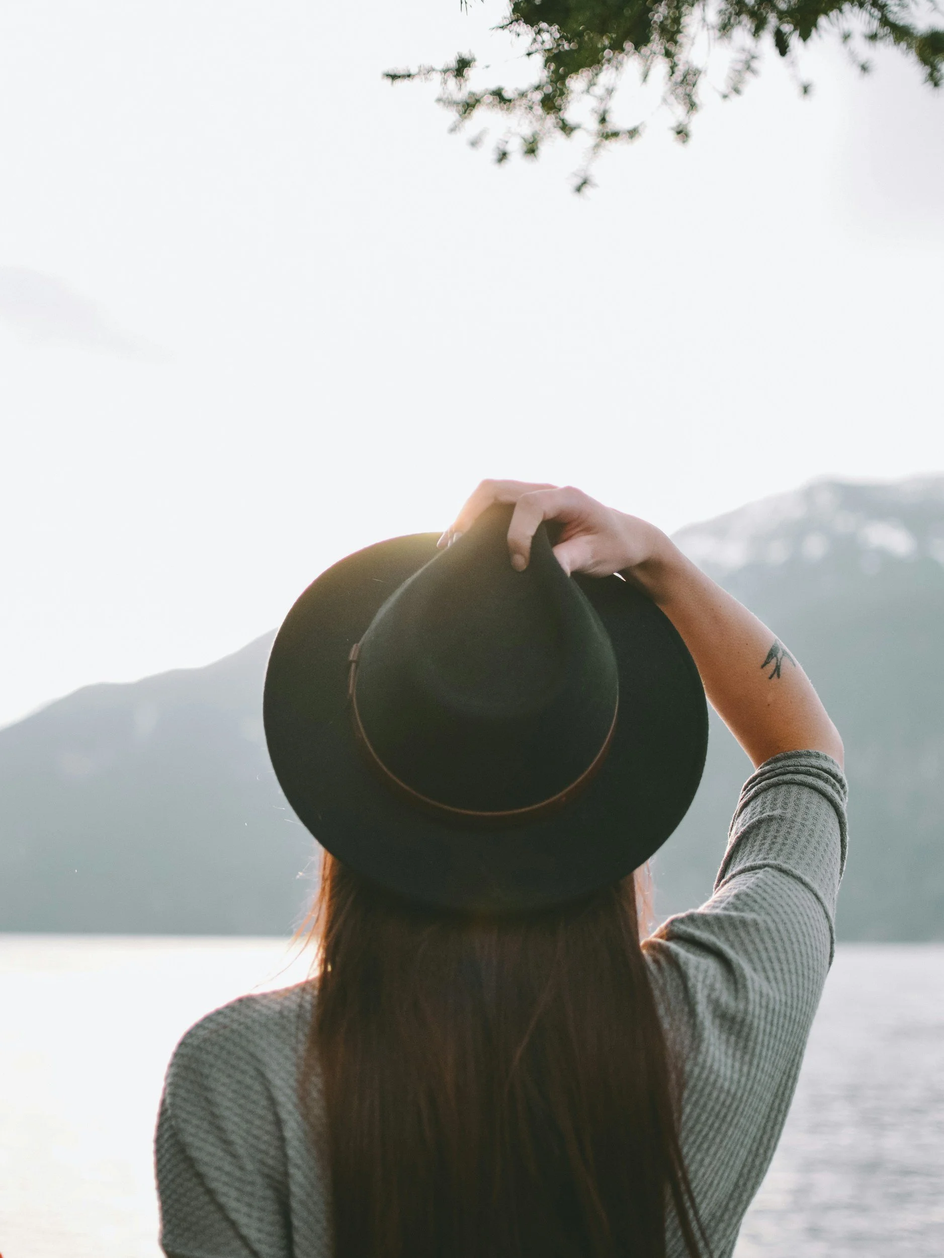 Person with long hair holding a black wide-brimmed hat on their head, facing a landscape with water and mountains, during daylight.