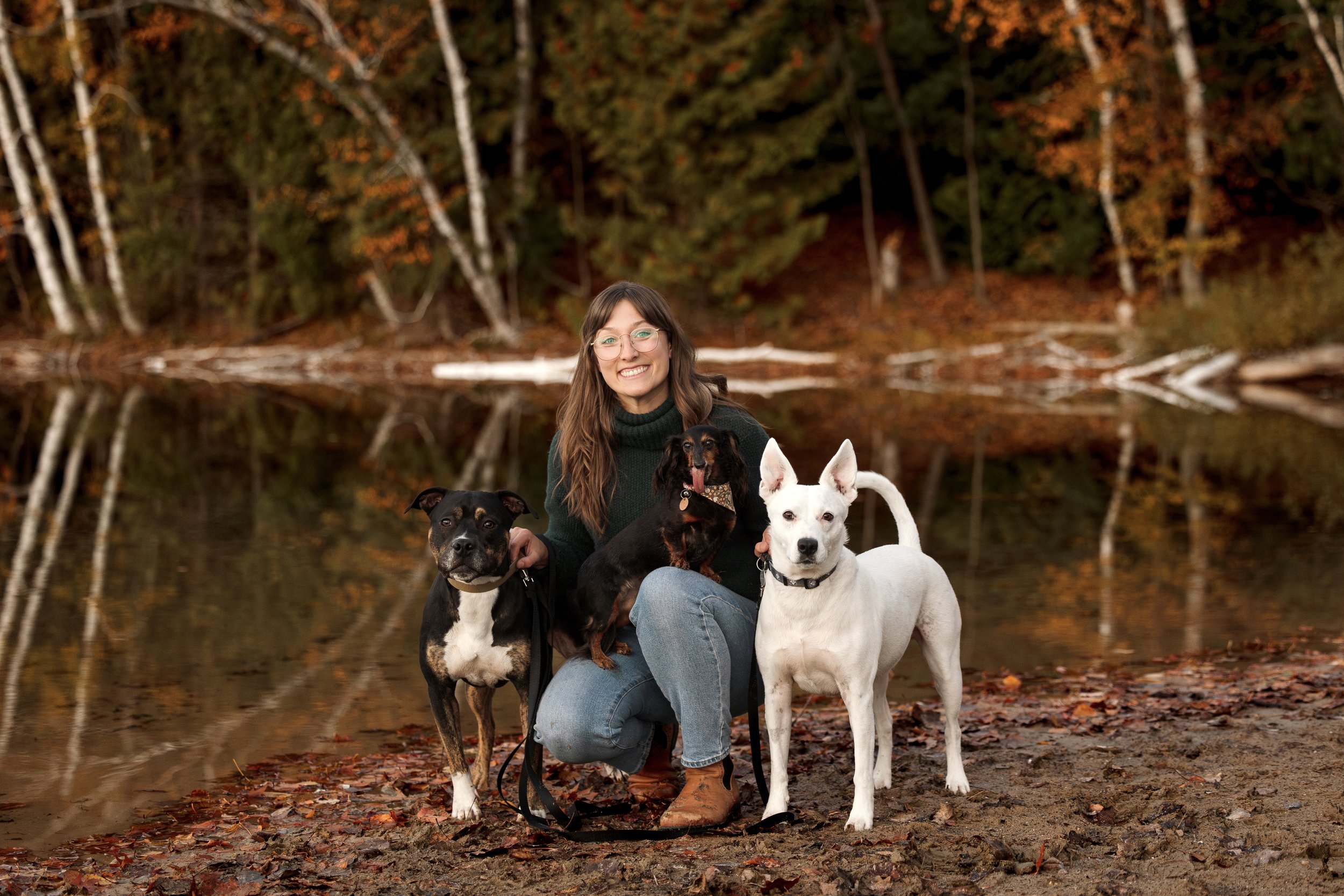 A woman with glasses and long brown hair, wearing a dark green sweater and jeans, smiling and kneeling on a leaf-covered ground next to three dogs near a calm body of water with an autumn forest background.