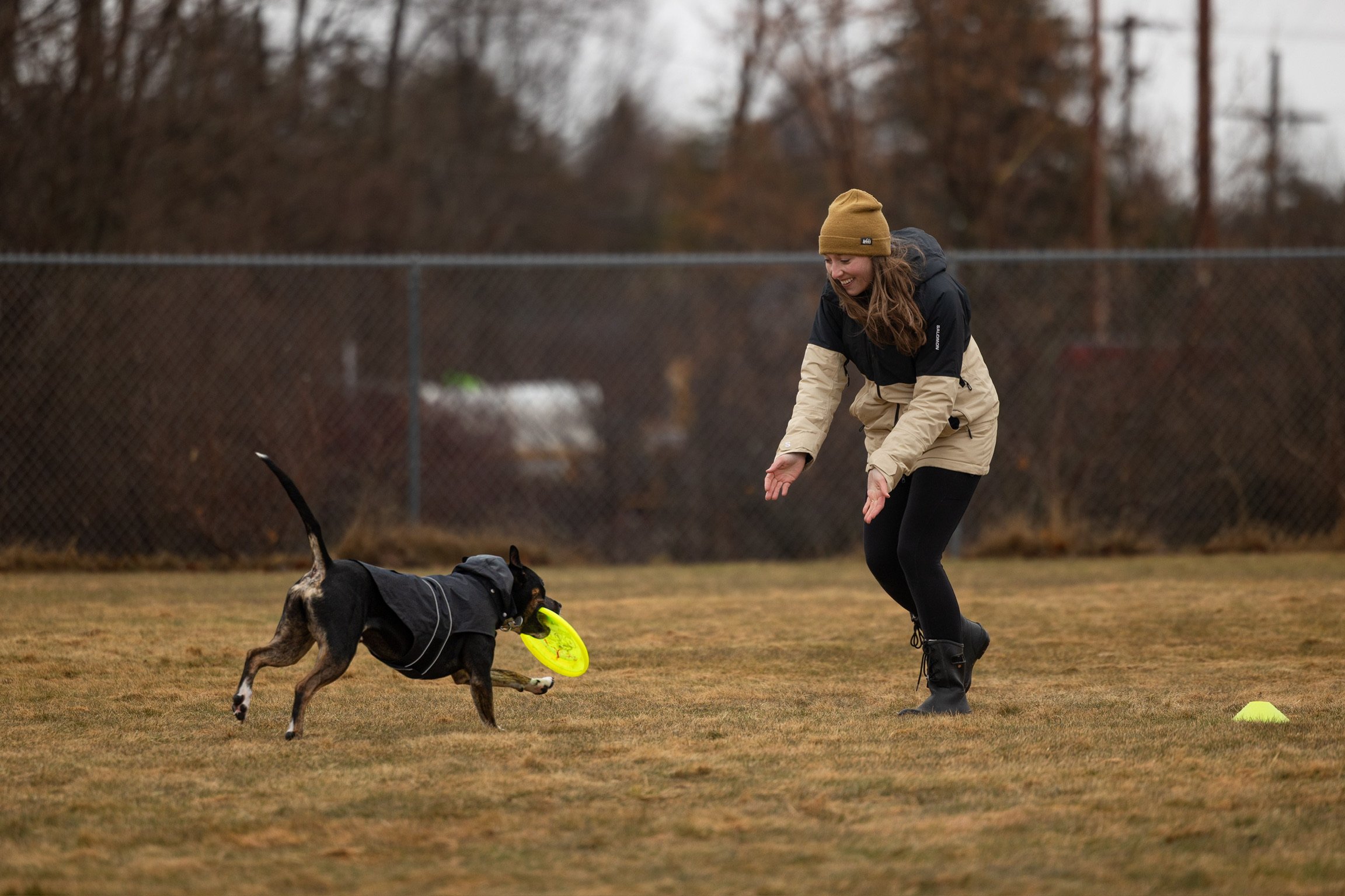 A woman playing fetch with a dog at a park on a cloudy day. The dog is catching a yellow frisbee, and the woman is smiling and dressed warmly in a beige and black jacket and a mustard-yellow beanie.