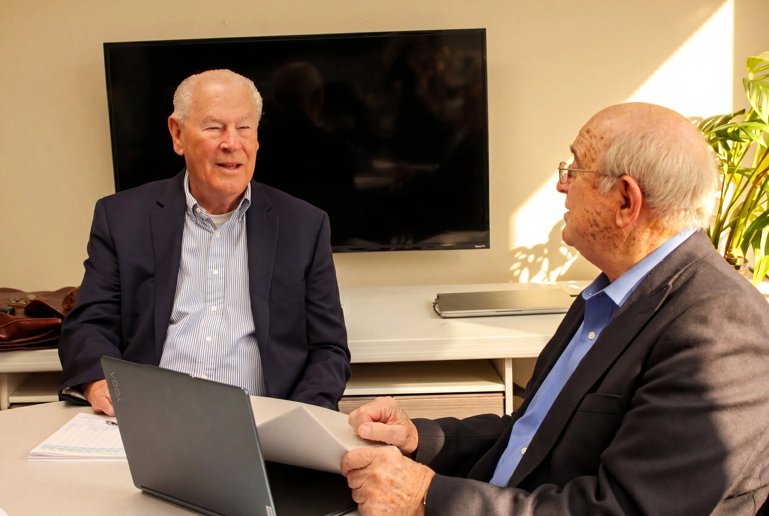 Fran DiBacco and Bill Bradley having a conversation in an office setting. Fran is seated with a laptop in front of him, and Bill is holding papers. They are engaged in a discussion, with a TV and a plant visible in the background