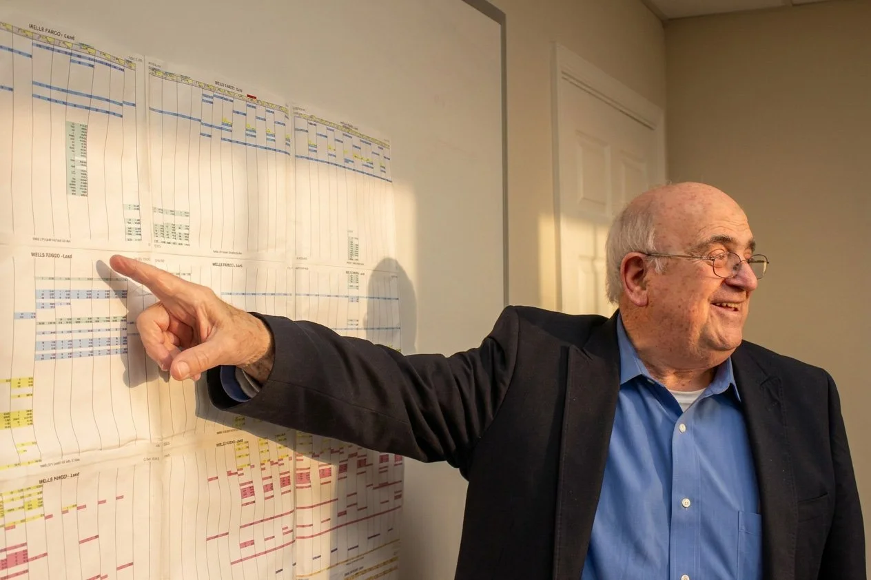 Fran DiBacco in a blue shirt and black blazer points at a large chart or schedule on a whiteboard, smiling and wearing glasses.