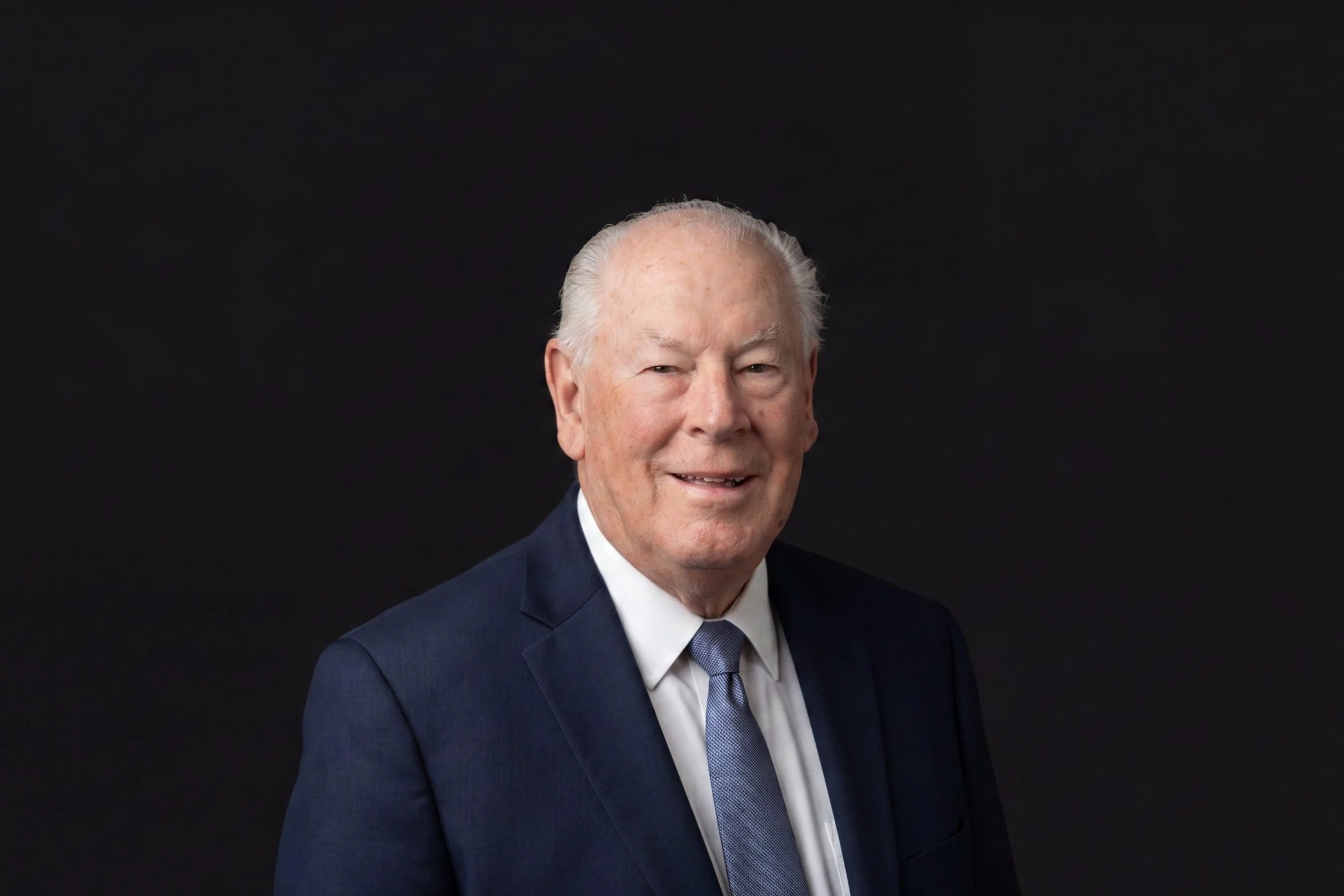 Portrait of Bill Bradley with white hair wearing a navy blue suit, white shirt, and tie, smiling against a black background.