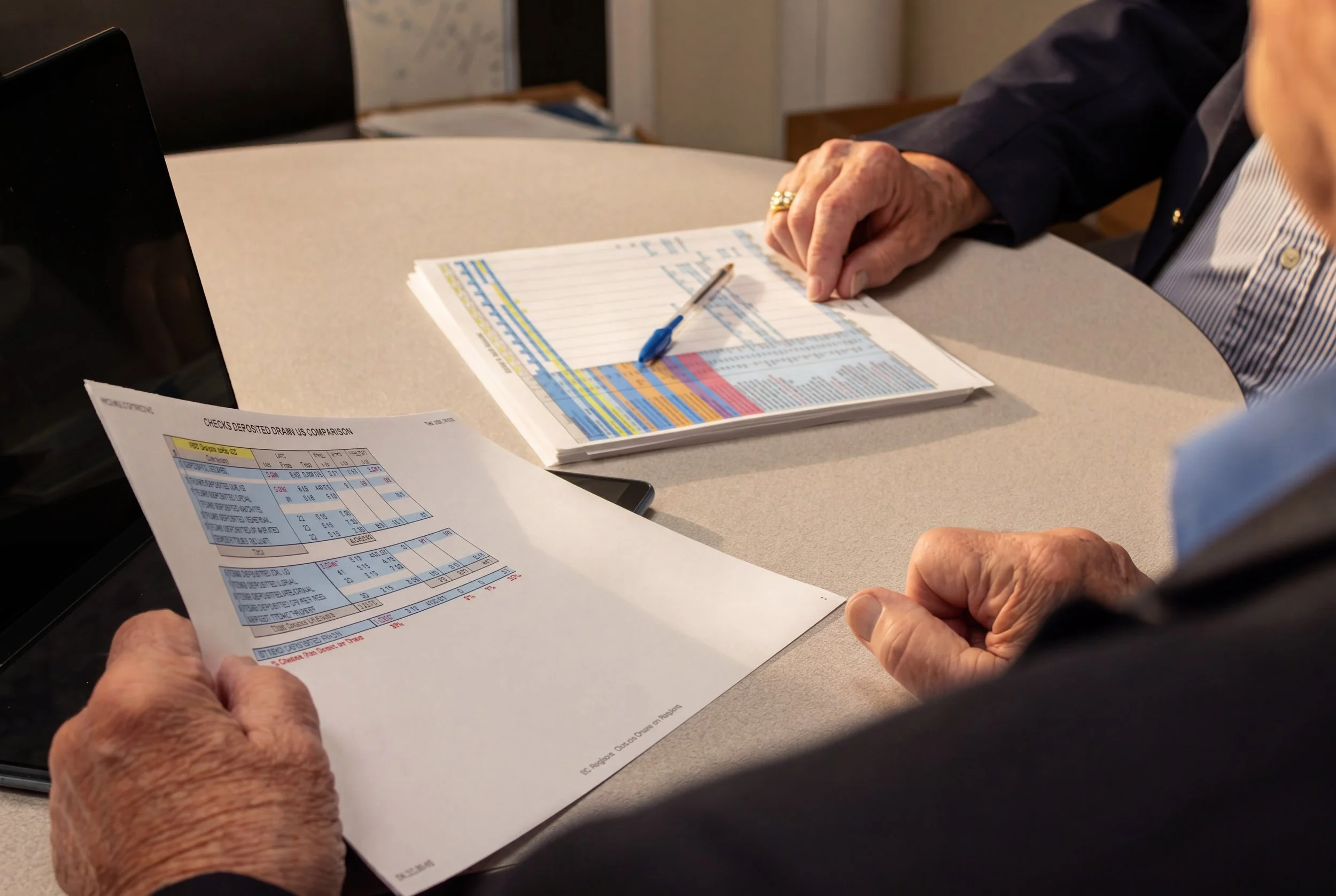Close-up of elderly person's hand holding a financial report, with another person pointing at the report and an open wallet on a table.