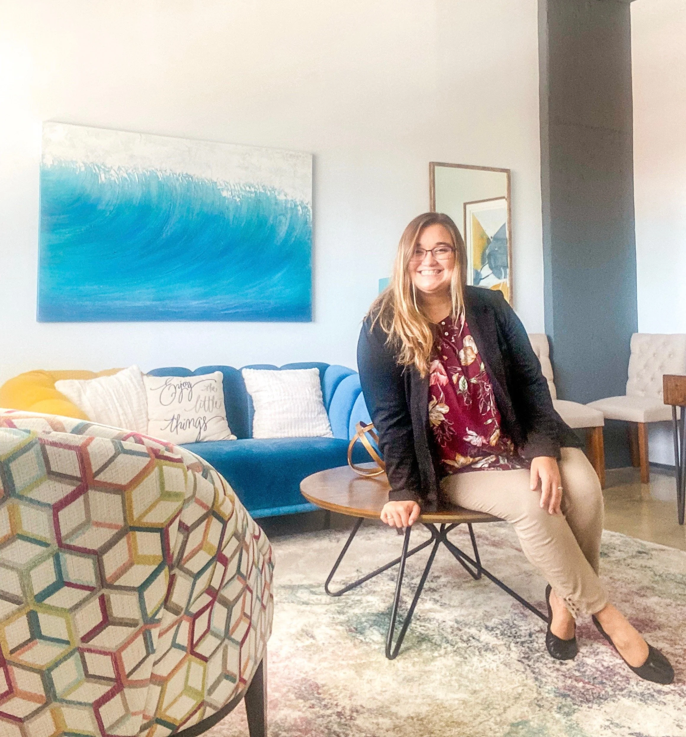 A woman sitting on a round wooden table in a living room, smiling, with a colorful patterned armchair in the foreground, and a blue sofa with decorative pillows, abstract artwork on the wall, and additional seating in the background.