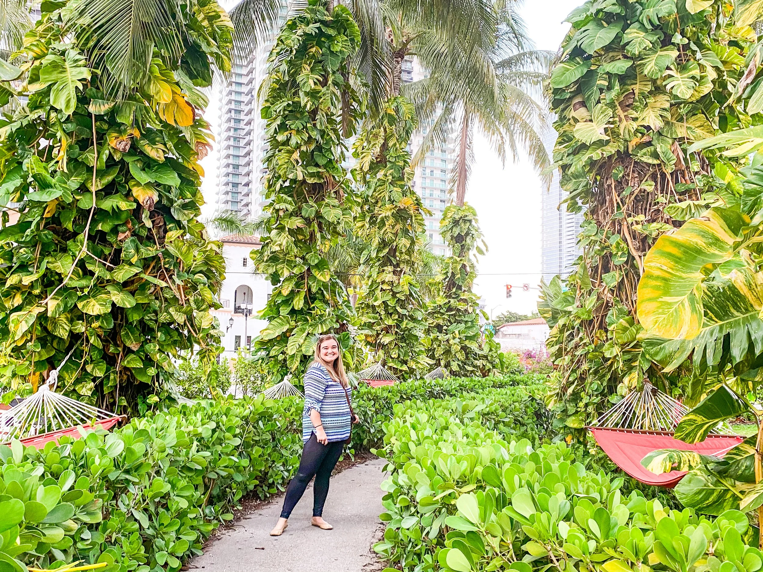 A smiling woman stands barefoot in a lush urban garden with tall green trees, dense bushes, and pink hammocks, with modern skyscrapers in the background.