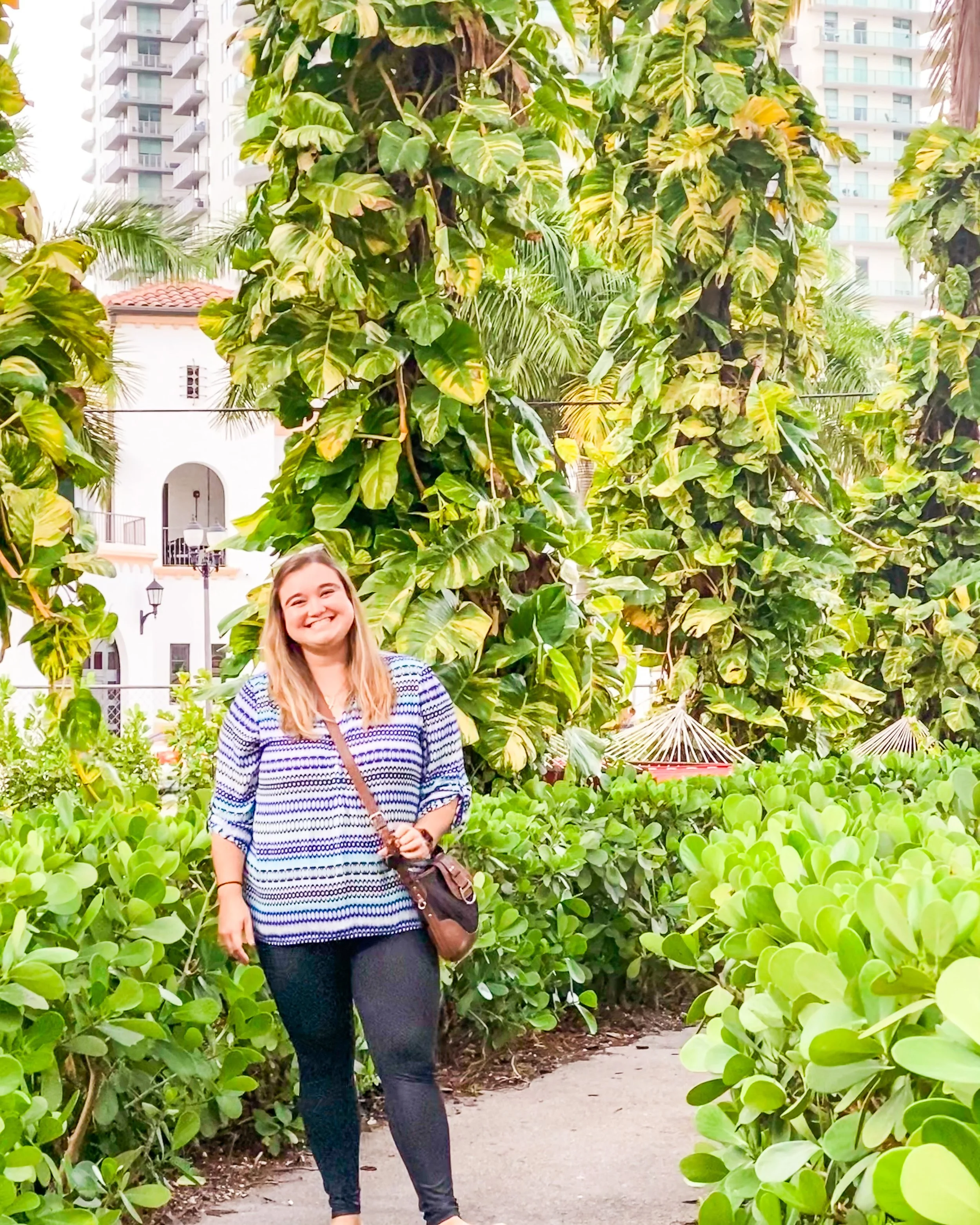A young woman with blonde hair smiling and standing on a garden path surrounded by lush green plants and large leafy trees.