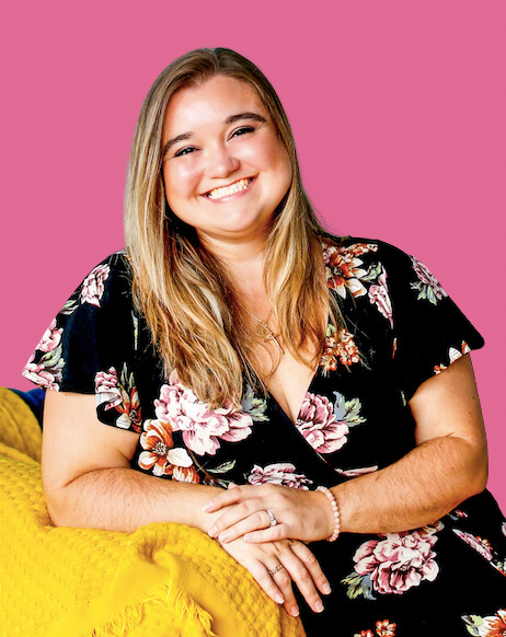A woman with long blonde hair smiling, wearing a black floral dress, sitting on a yellow textured couch against a pink background.