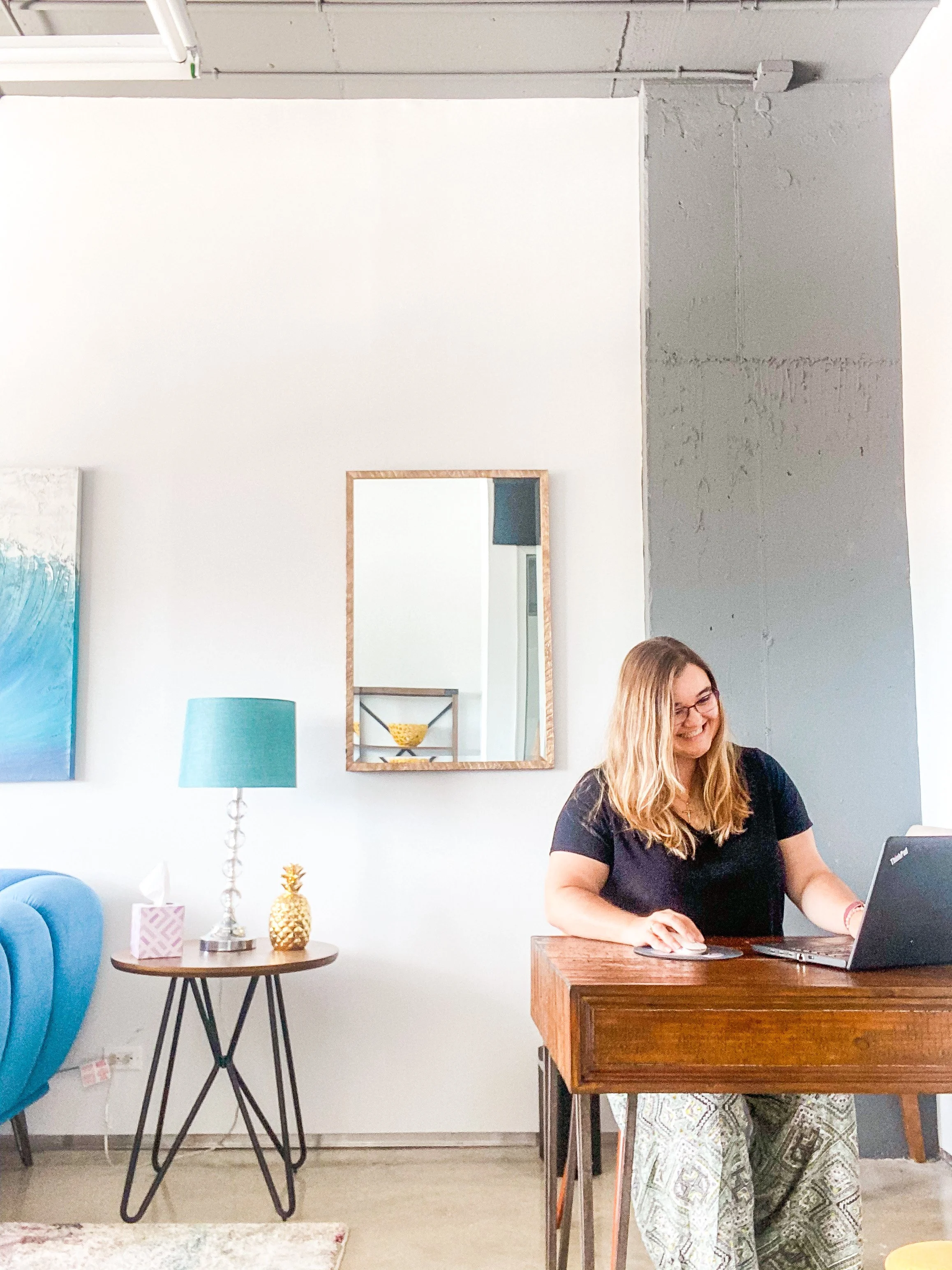 A woman with glasses and long hair working on a laptop at a wooden desk in a brightly decorated room.