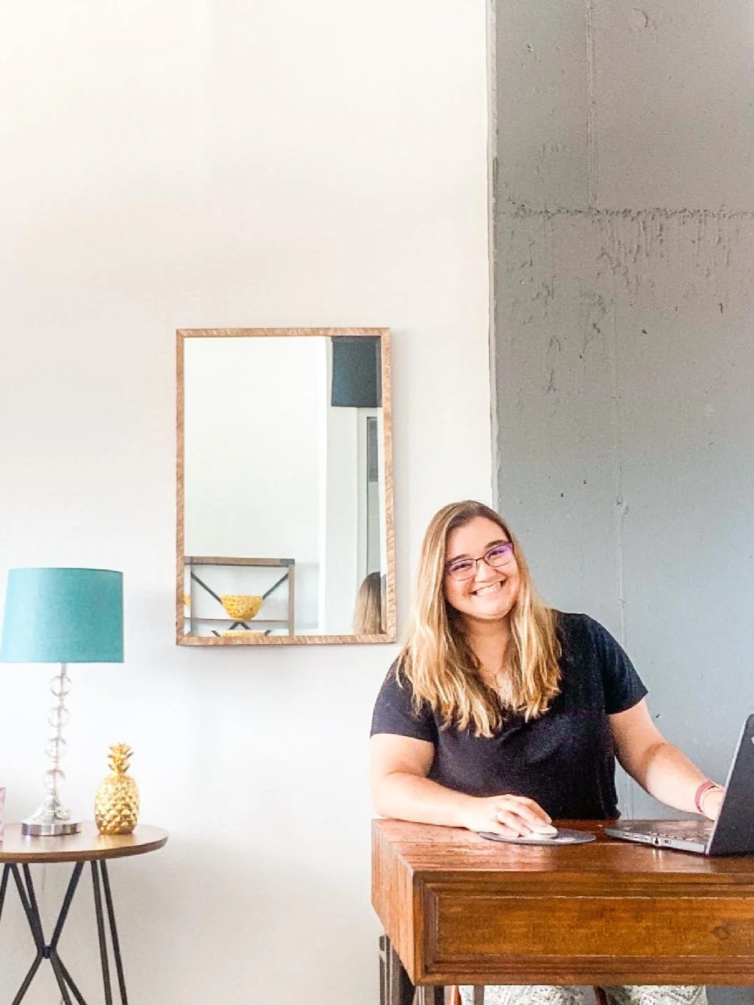 A smiling woman with glasses working on a laptop at a wooden desk in a modern, minimalistic room with a mirror, a teal lamp, and a decorative pineapple on a side table.
