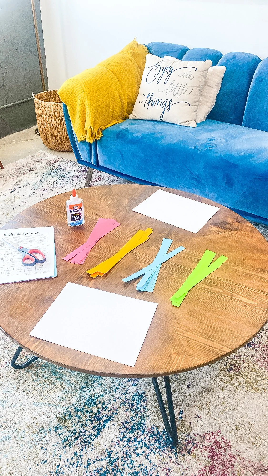 A round wooden table with colorful paper strips, blank sheets, a bottle of glue, scissors, and a worksheet, set in a living room with a blue sofa, yellow blanket, decorative pillows, a basket, and a multicolored rug.