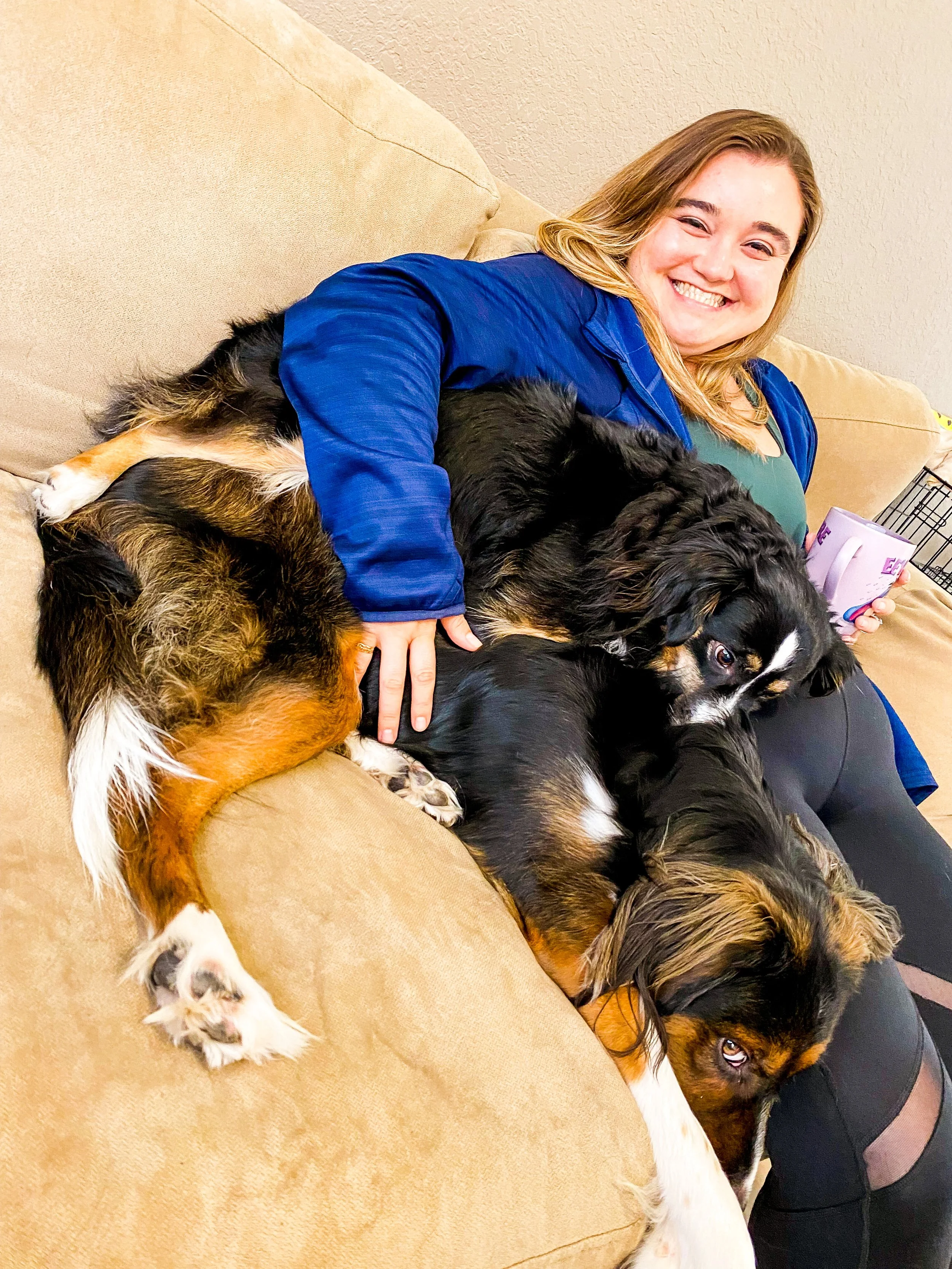 A woman with blonde hair wearing a blue jacket is sitting on a beige couch, smiling happily, while cuddling two large black, brown, and white dogs.