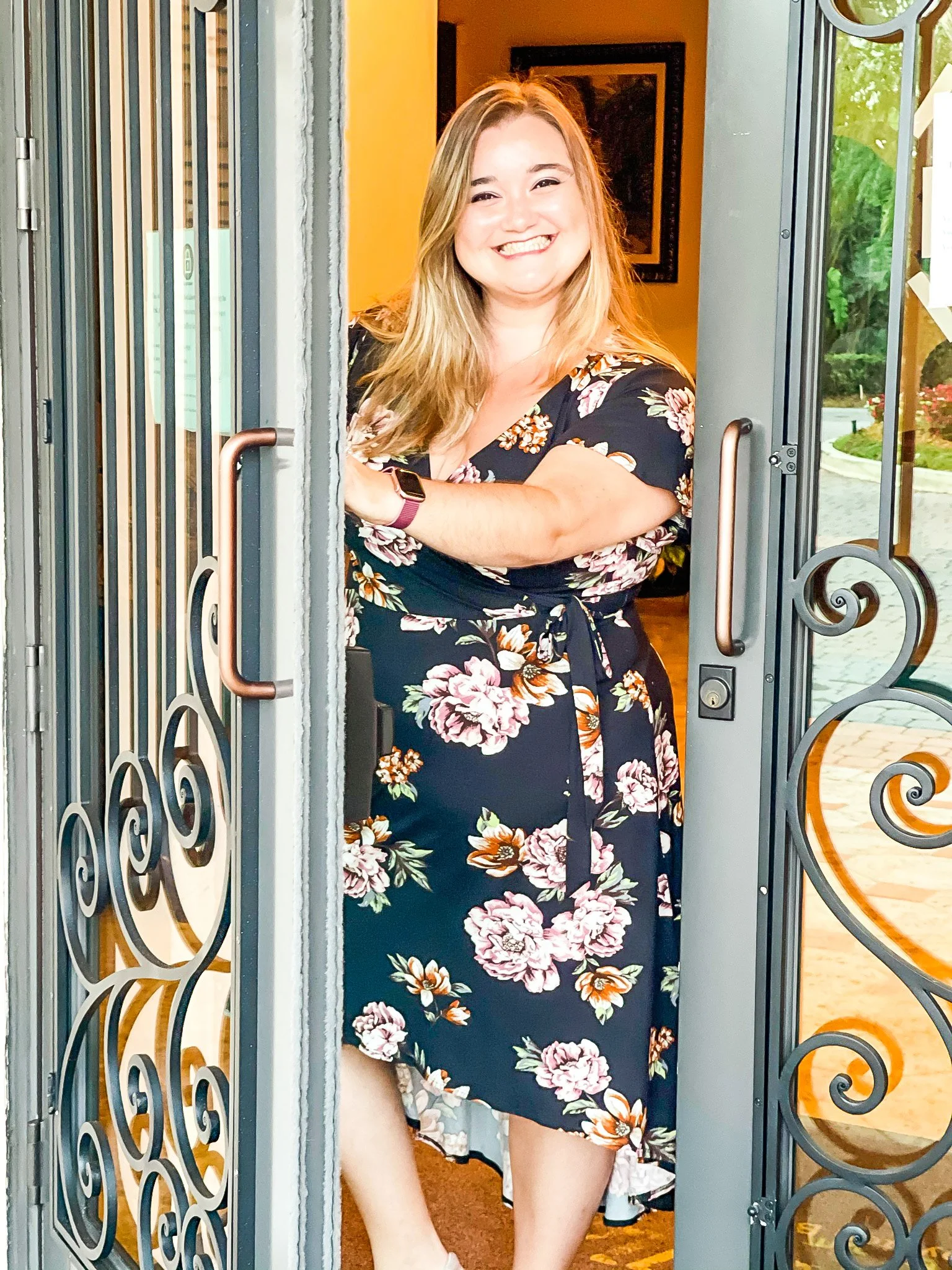 A woman with long blonde hair in a floral dress smiling and leaning on a doorframe, with greenery visible outside.