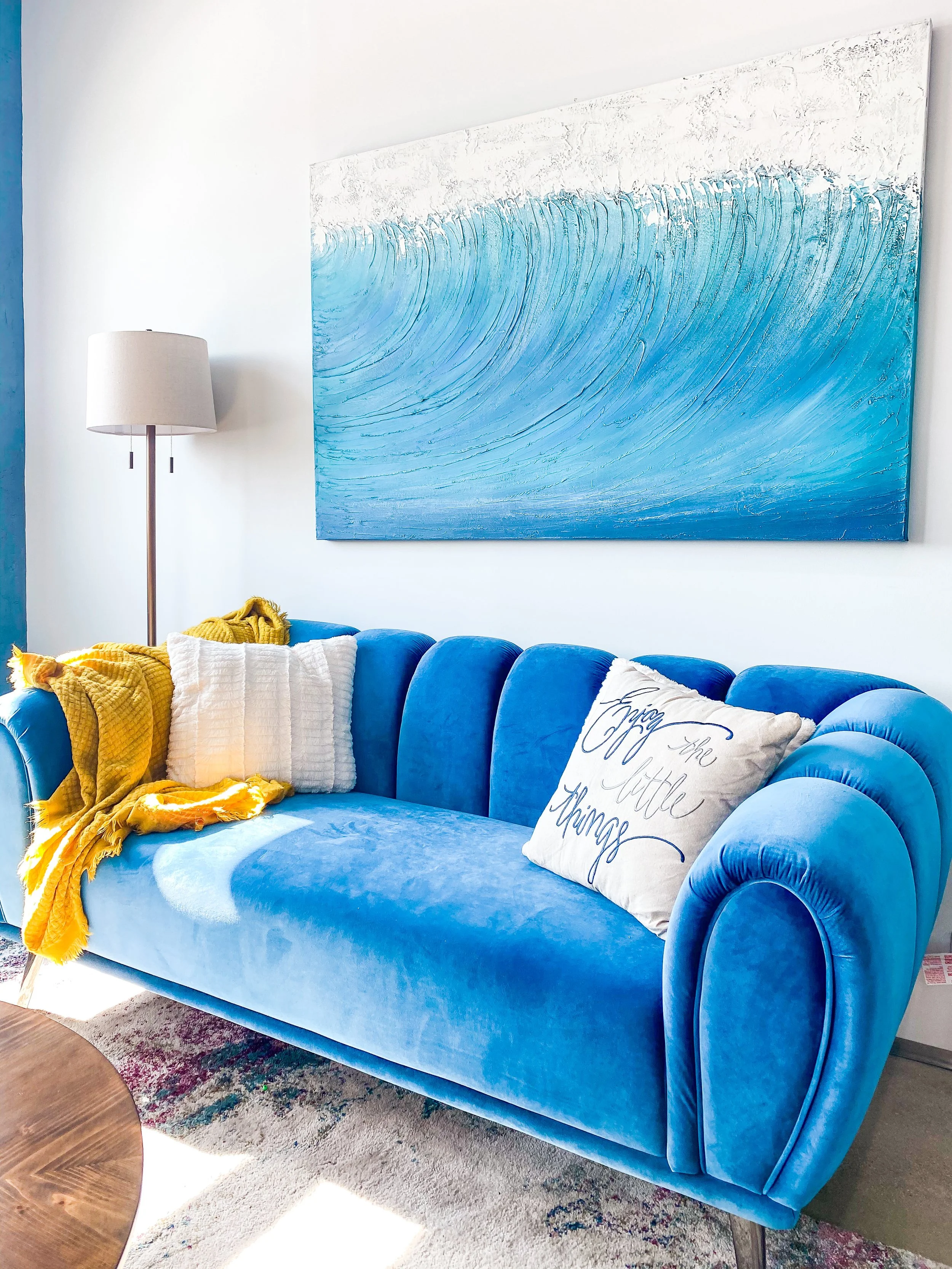 A bright blue velvet sofa with rolled arms and decorative pillows, including a white one with black cursive writing that says 'enjoy the little things', in a living room with a large abstract blue and white wall art and a floor lamp.