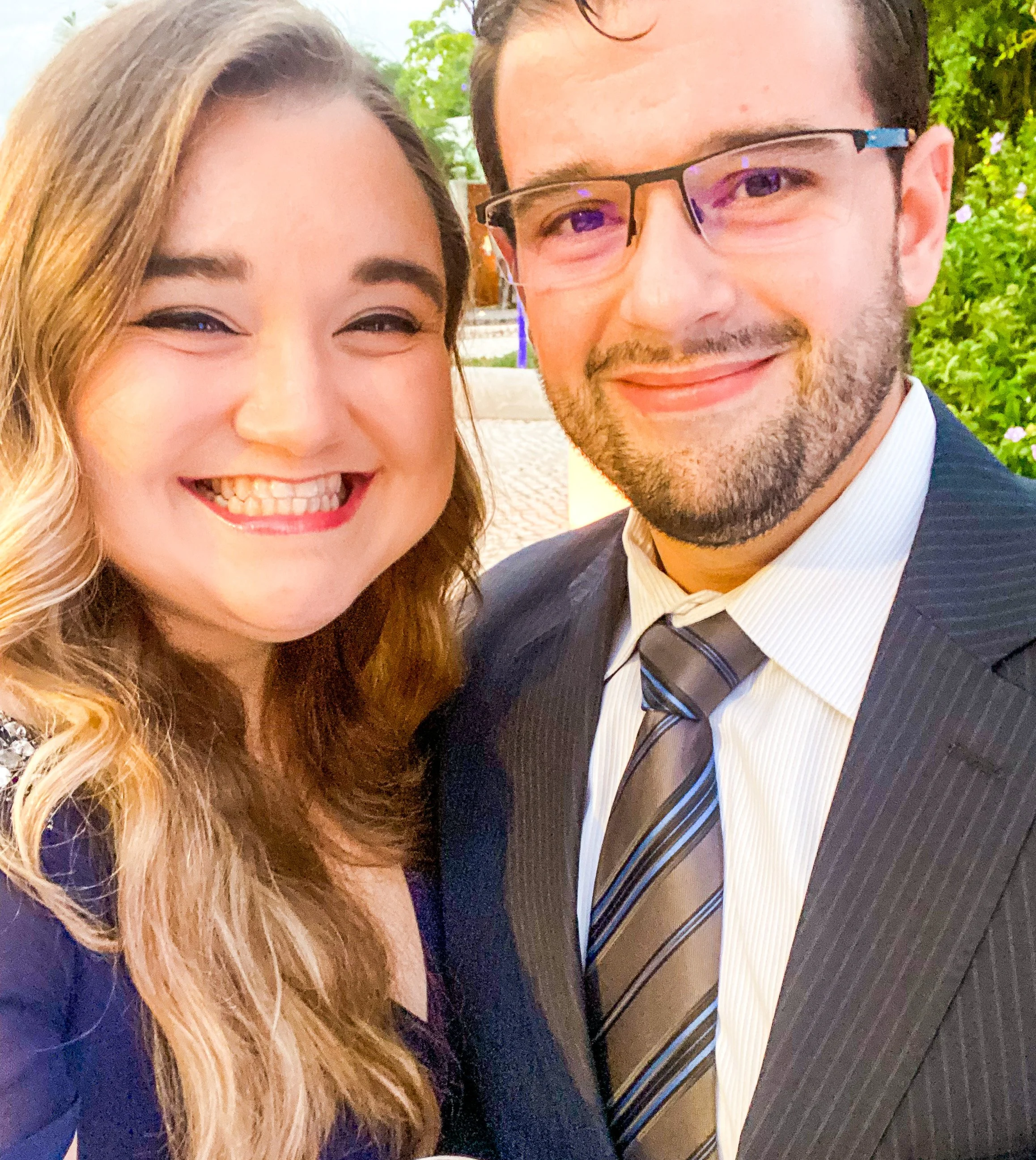 A smiling woman with long, wavy blonde hair and a man with short dark hair, glasses, and a beard, dressed in a suit and tie, standing outdoors with greenery in the background.