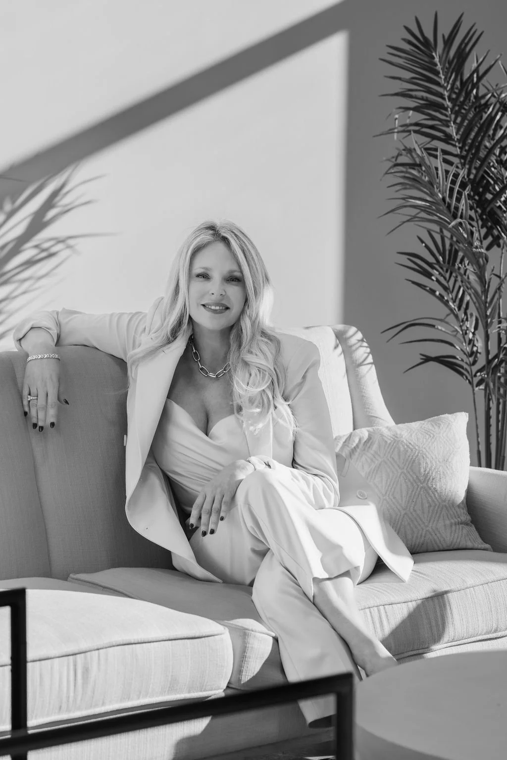 Black and white photo of a smiling woman sitting on a sofa, wearing a light-colored suit and jewelry, with plants in the background and sunlight casting shadows.