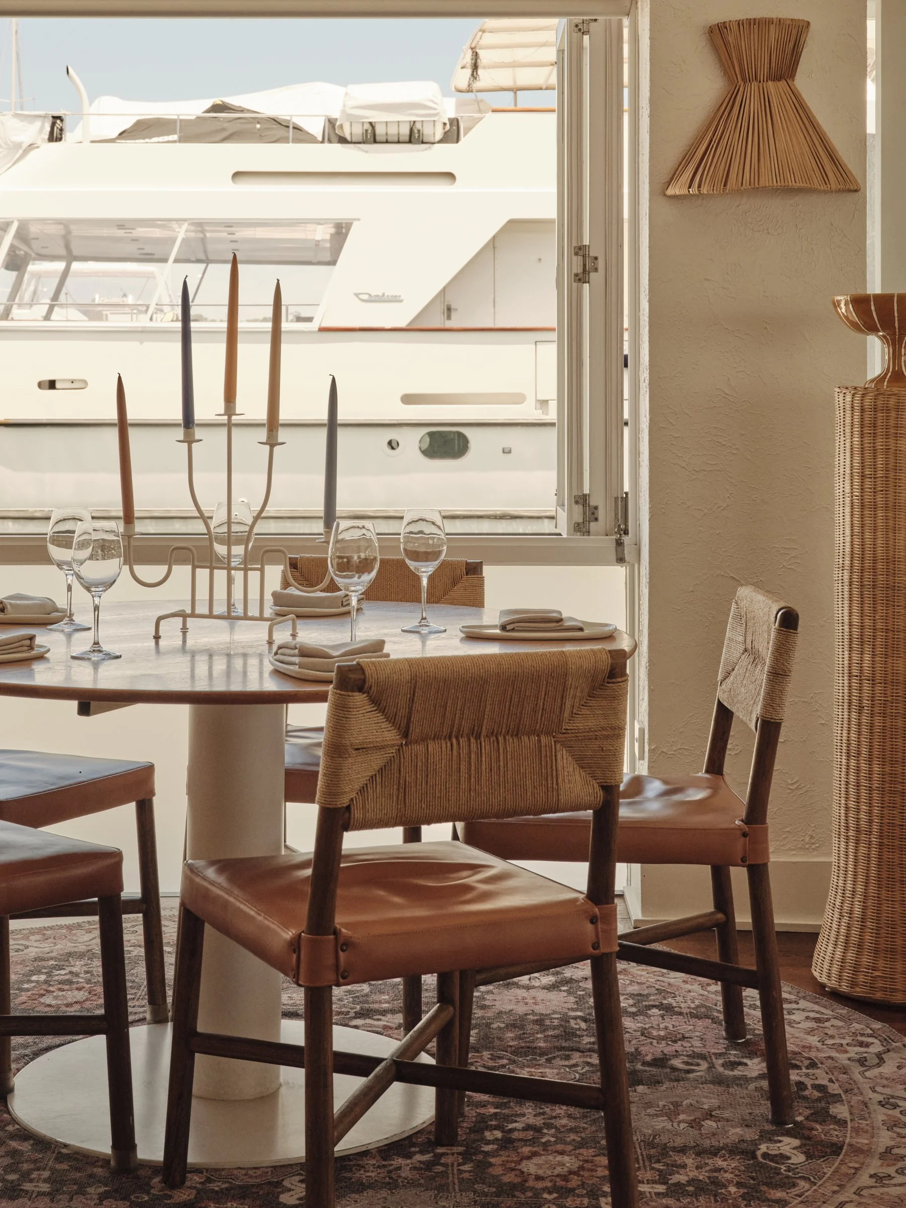 A dining table set for four near a large window overlooking a marina with boats. The table has a candleholder with five colorful candles, wine glasses, and neatly folded napkins. The room features wicker and wooden chairs, a patterned rug, and decorative wicker wall art.