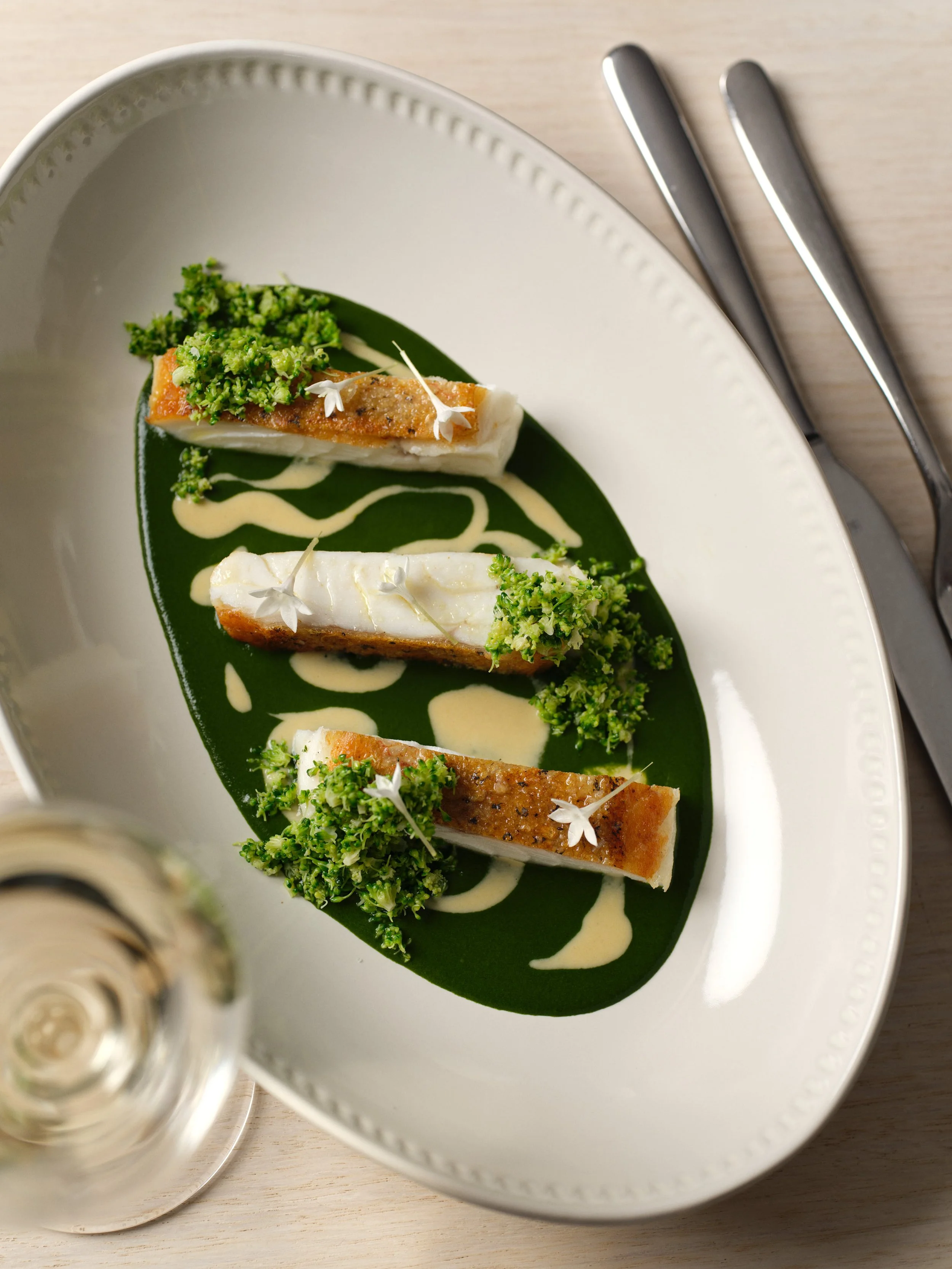 A gourmet dish featuring three pieces of cooked fish with crispy skin, garnished with broccoli and edible white flowers, served on a white plate with a green sauce and white drizzle, accompanied by silverware and a glass of white wine.