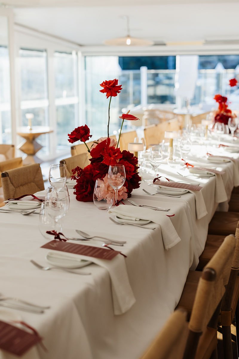 Elegant dining table set for a special event with white tablecloths, red floral centerpieces, wine glasses, plates, and utensils in a bright, modern room.