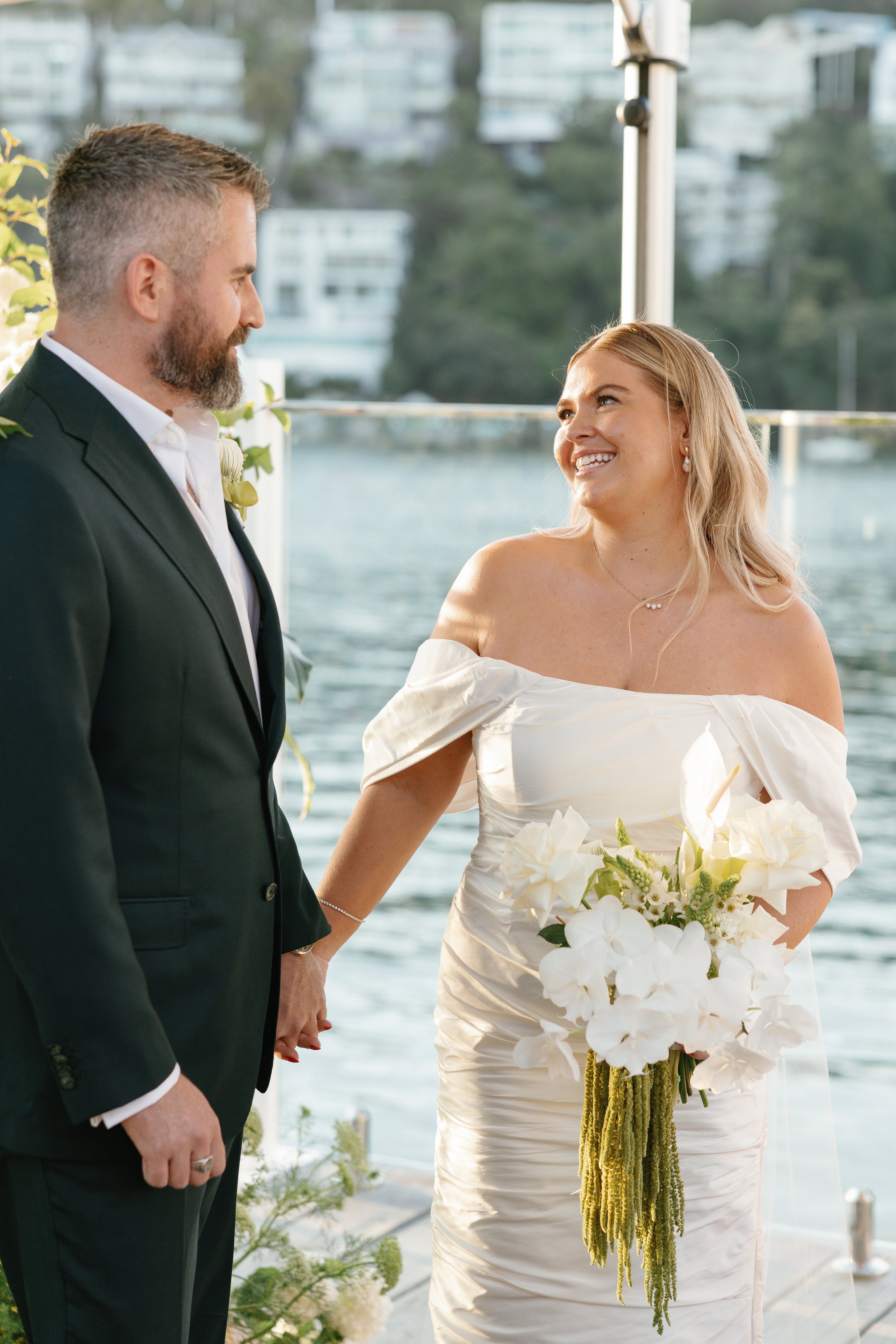 A bride and groom holding hands and smiling at each other during their wedding ceremony by a waterfront with modern buildings in the background.