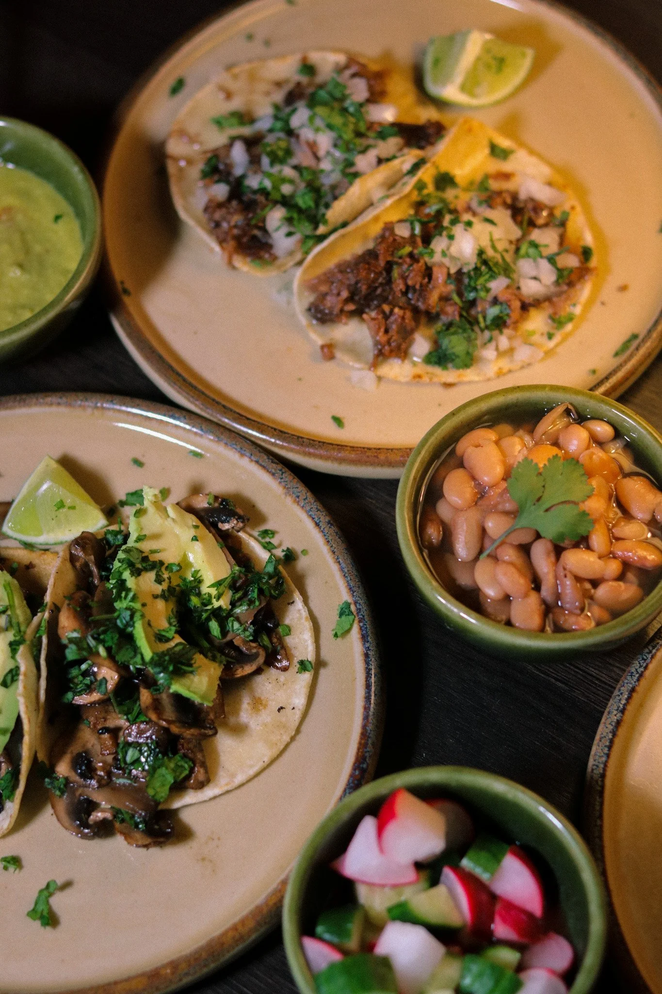 Assorted Mexican dishes including tacos with meat and cilantro, served with lime wedges, salsa, beans, and pickled vegetables on ceramic plates and bowls.