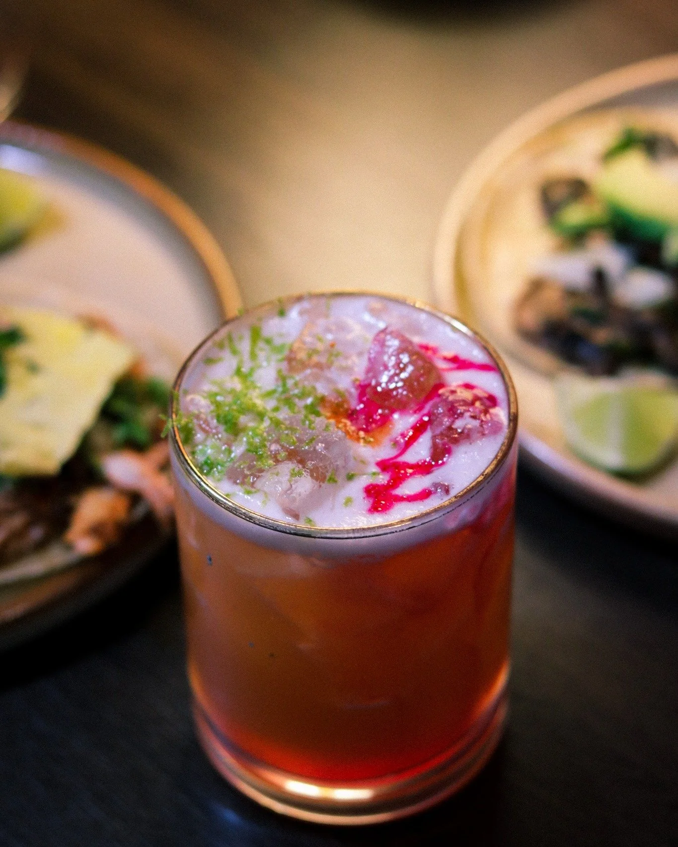 Look closely &mdash; that's the detail that separates a drink from a cocktail.
Crushed ice, a ribbon of deep red bleeding through, fresh lime zest grated right over the top.
Nothing on this glass got there by accident. Every garnish is a decision.
Ta