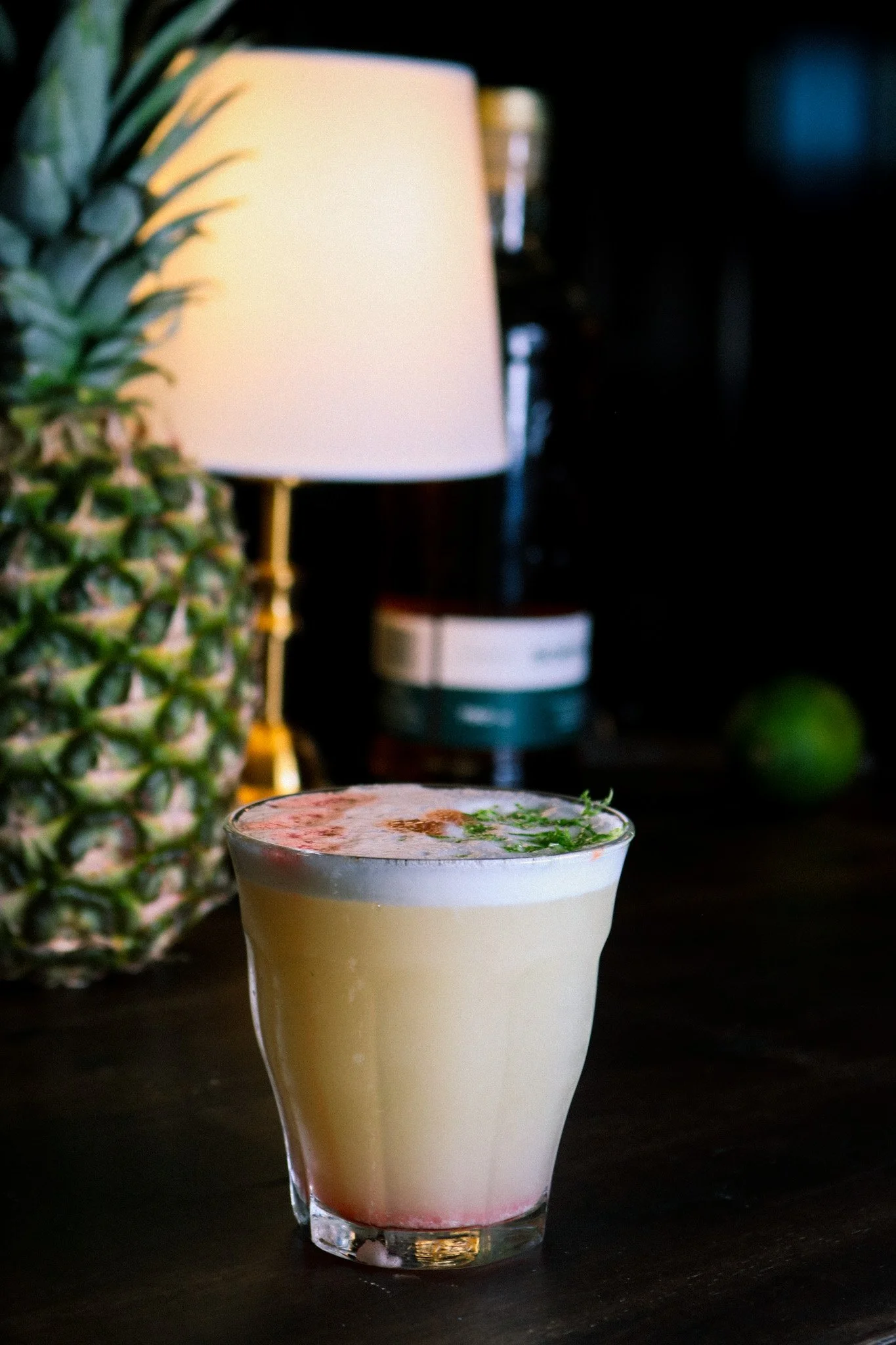 A layered cocktail drink garnished with herbs, placed on a dark surface with a pineapple and a lamp in the background.