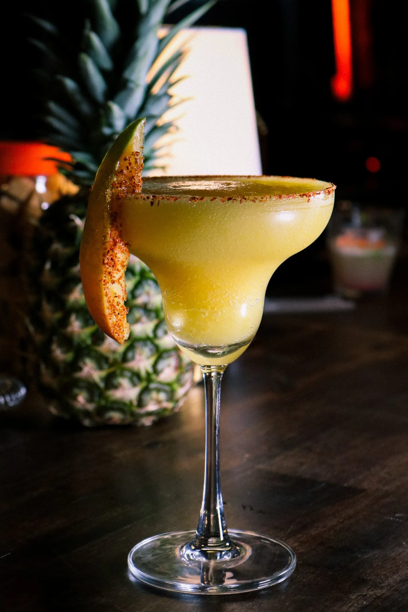 A yellow margarita cocktail in a glass with a salted rim, garnished with a lemon wedge and chili powder, with a pineapple and a blurred background.