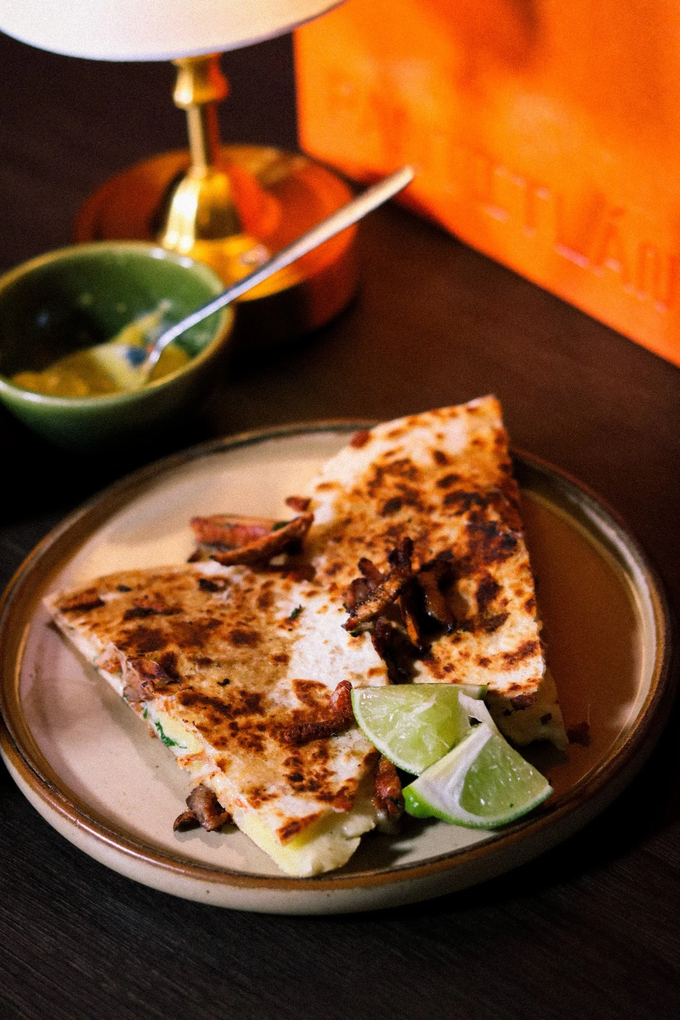 Grilled quesadilla with lime wedges on a ceramic plate, with a small green bowl of salsa and a white lamp in the background.