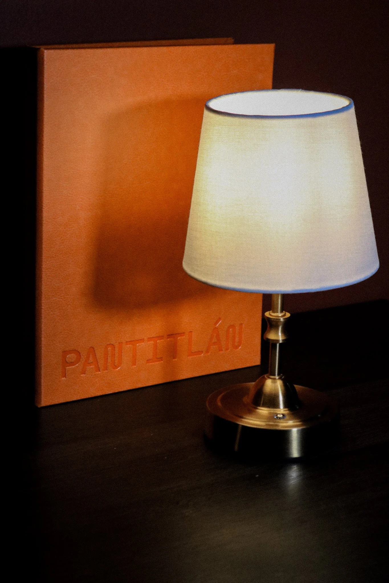 A table lamp with a white shade on a dark surface, next to an orange box with the word 'PAUTITLÁU' embossed on it.