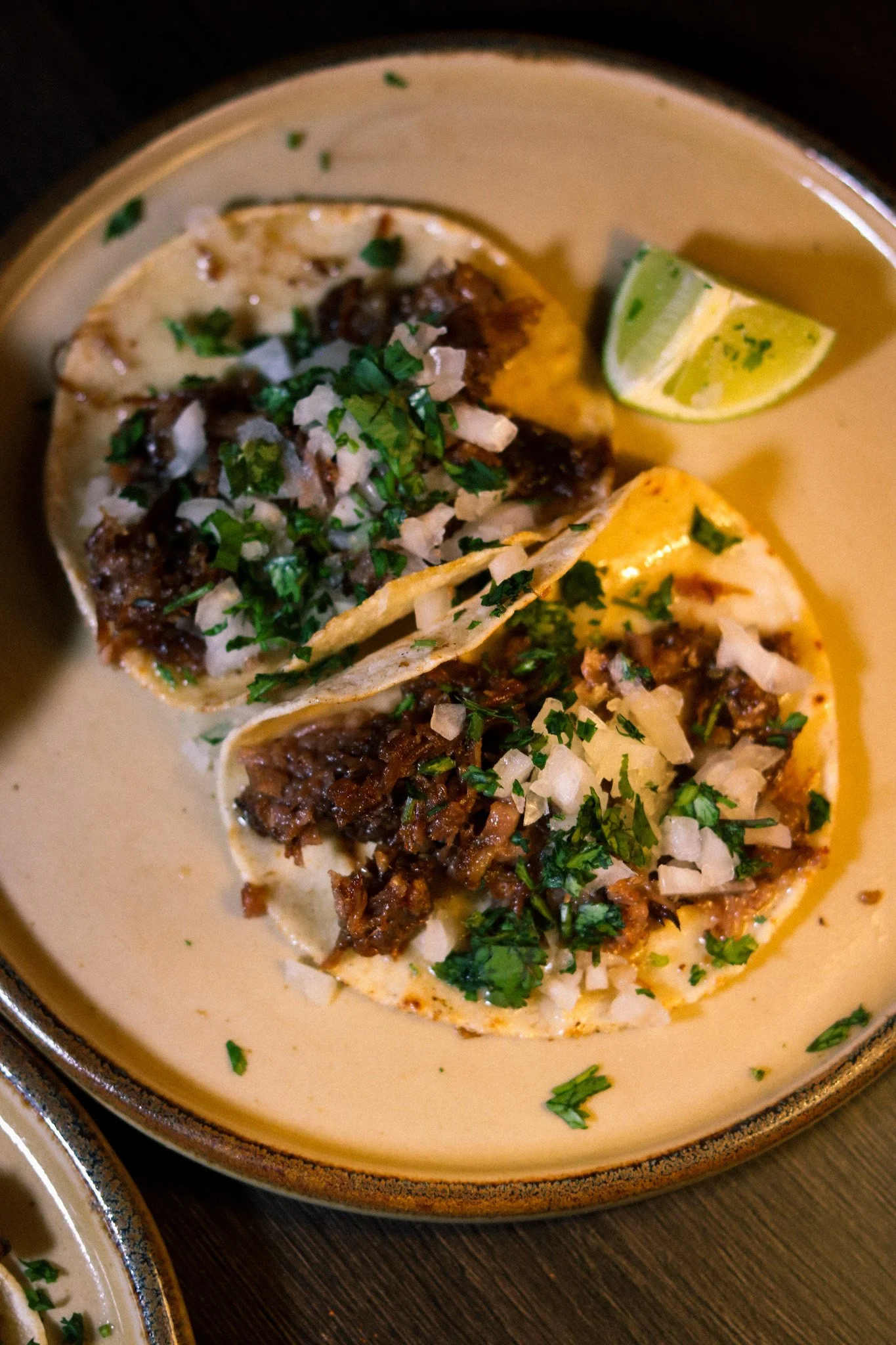 Three tacos with chopped onions and cilantro on a cream sauce, served with lime wedges on a beige plate.