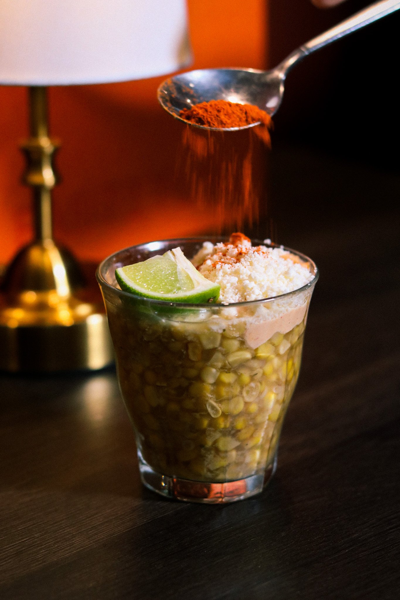 A glass filled with corn kernels, topped with shredded cheese, a lime wedge, and red chili powder being sprinkled from a spoon.