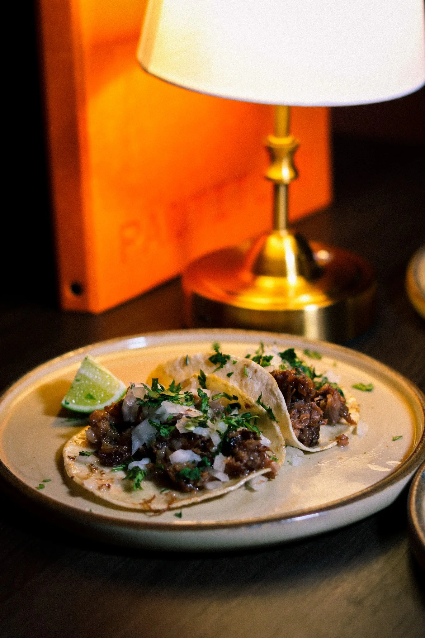 Three tacos filled with beef, topped with chopped onions, cilantro, and a lime wedge, served on a beige plate with a gold rim, placed on a dark table next to a brass lamp with a white shade.