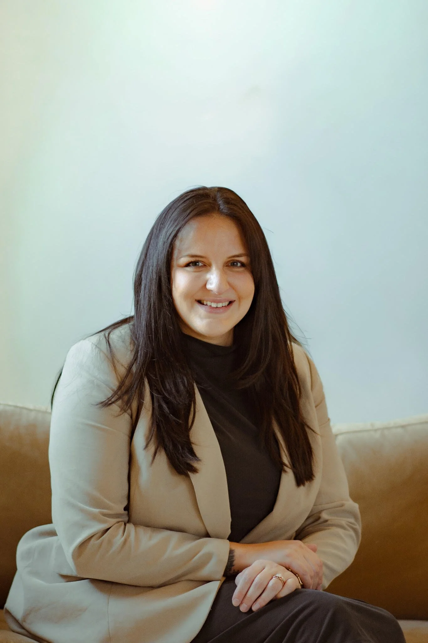 A woman with long dark hair, wearing a beige blazer and black shirt, sitting on a sofa and smiling.