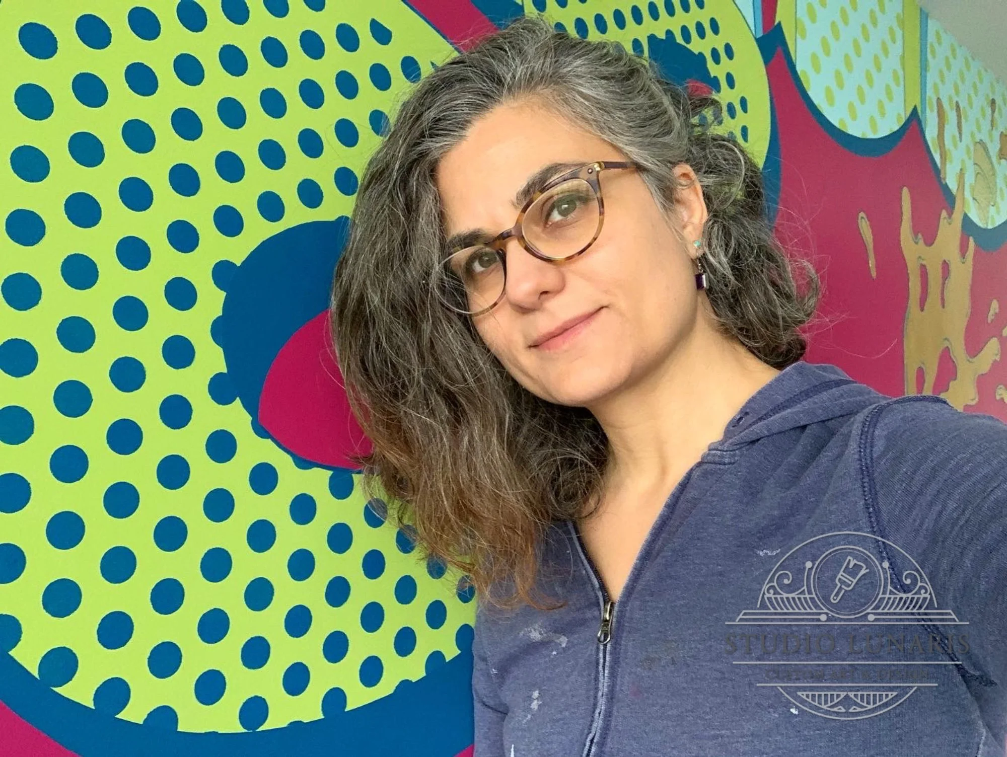 The artist with curly, shoulder-length gray hair wearing glasses and a navy hoodie taking a selfie in front of a colorful, abstract mural with yellow, blue, and pink patterns.