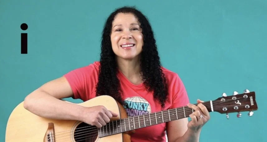 A woman with curly black hair wearing a pink T-shirt playing an acoustic guitar against a teal background.