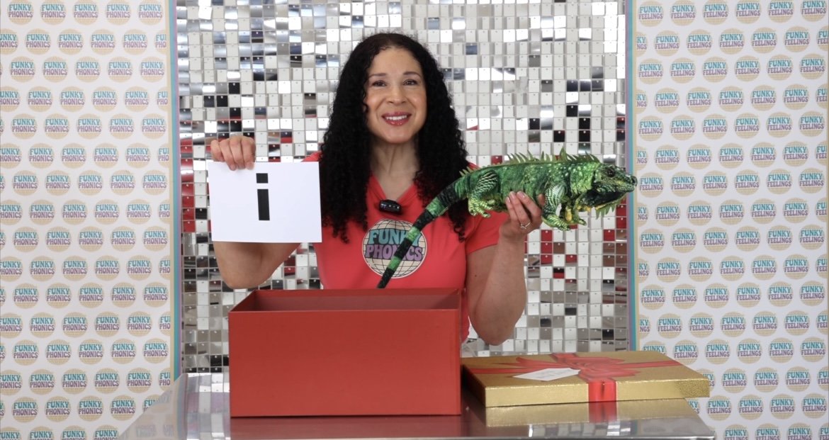 Woman with curly black hair smiling, holding a green chameleon and a card with the letter 'i', standing behind red and gold gift boxes, with a colorful mosaic background and a wall with a repeating 'FUNKY PHONICS' logo.
